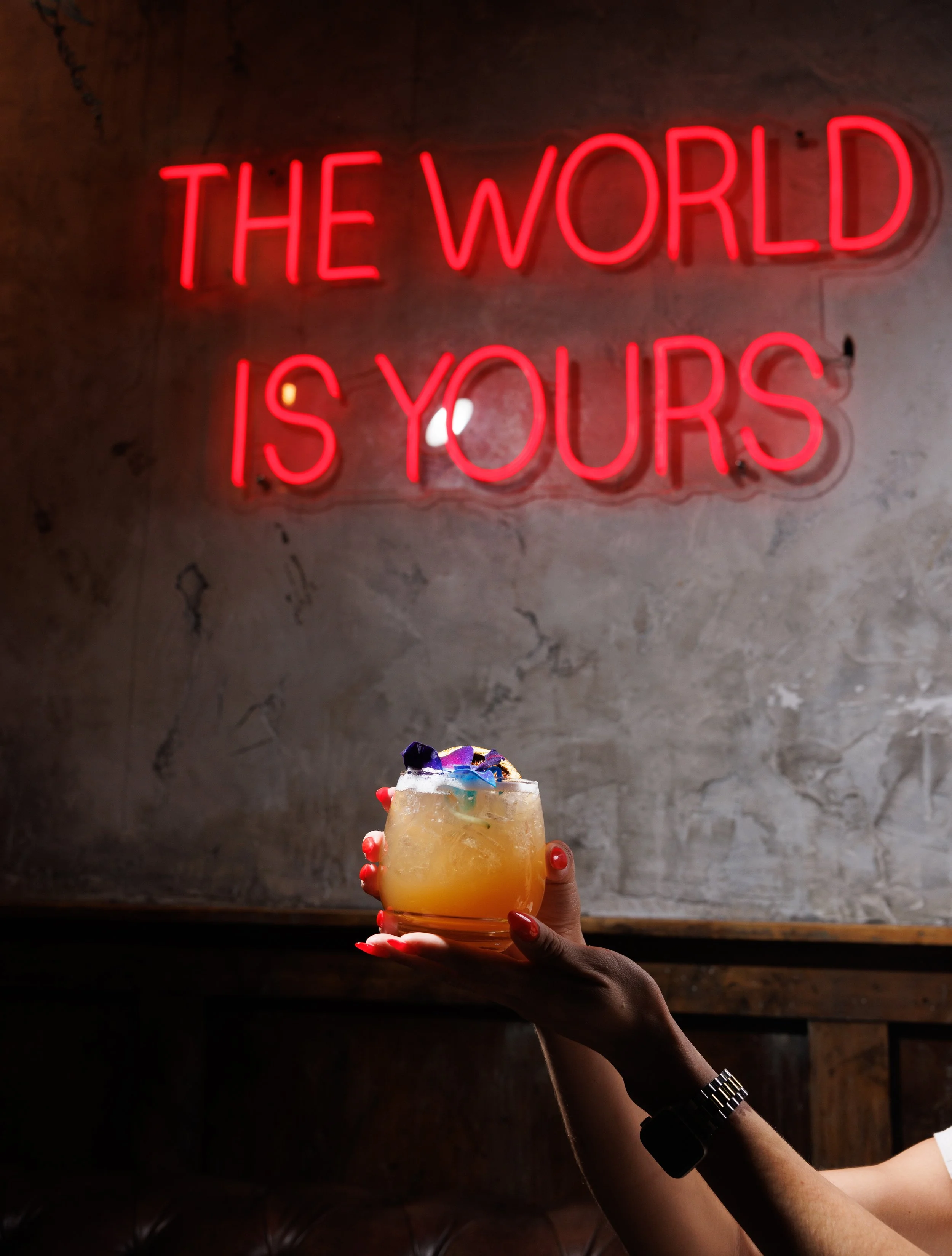 A person with painted red nails holding a tropical cocktail with colorful garnishes in front of a neon sign that says 'THE WORLD IS YOURS' in red letters.