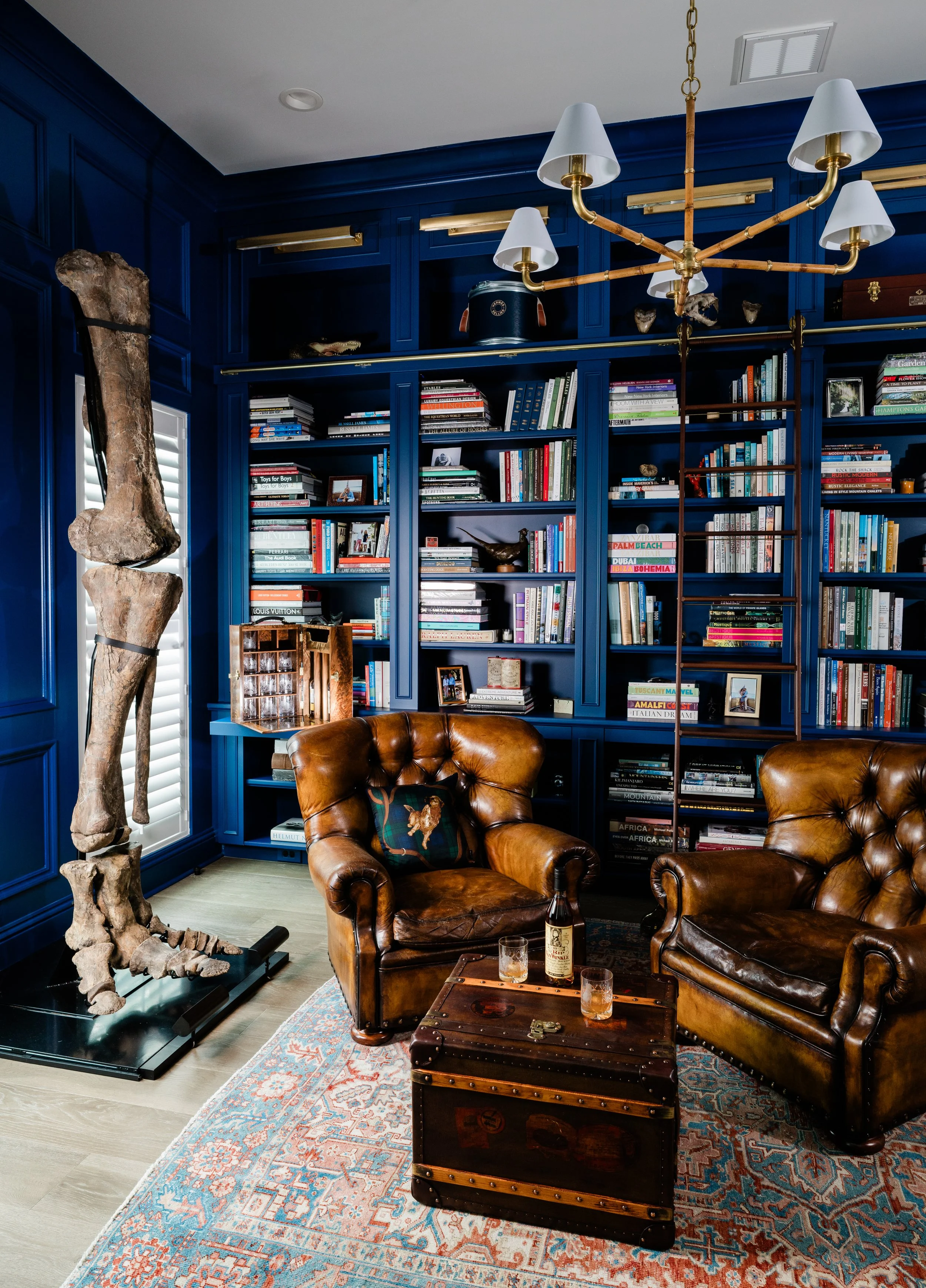 A cozy reading nook or living area with deep blue built-in bookshelves filled with books, framed photos, and decorative items. Two brown leather armchairs with tufted backs and rolled arms sit on a patterned rug. A wooden trunk with metal accents and