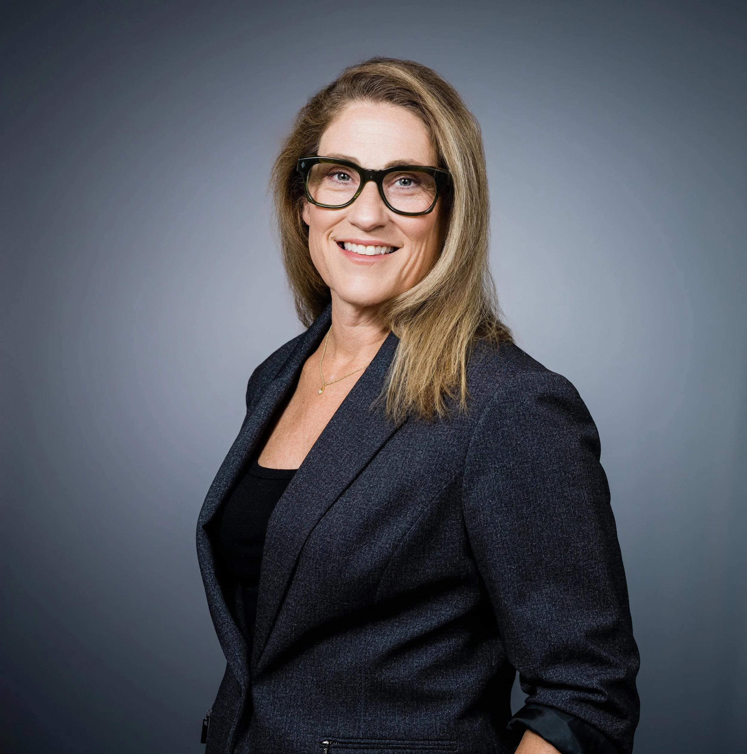 Portrait of a woman with long blonde hair wearing black-rimmed glasses, a dark blazer, and a black top, smiling against a plain gray background.