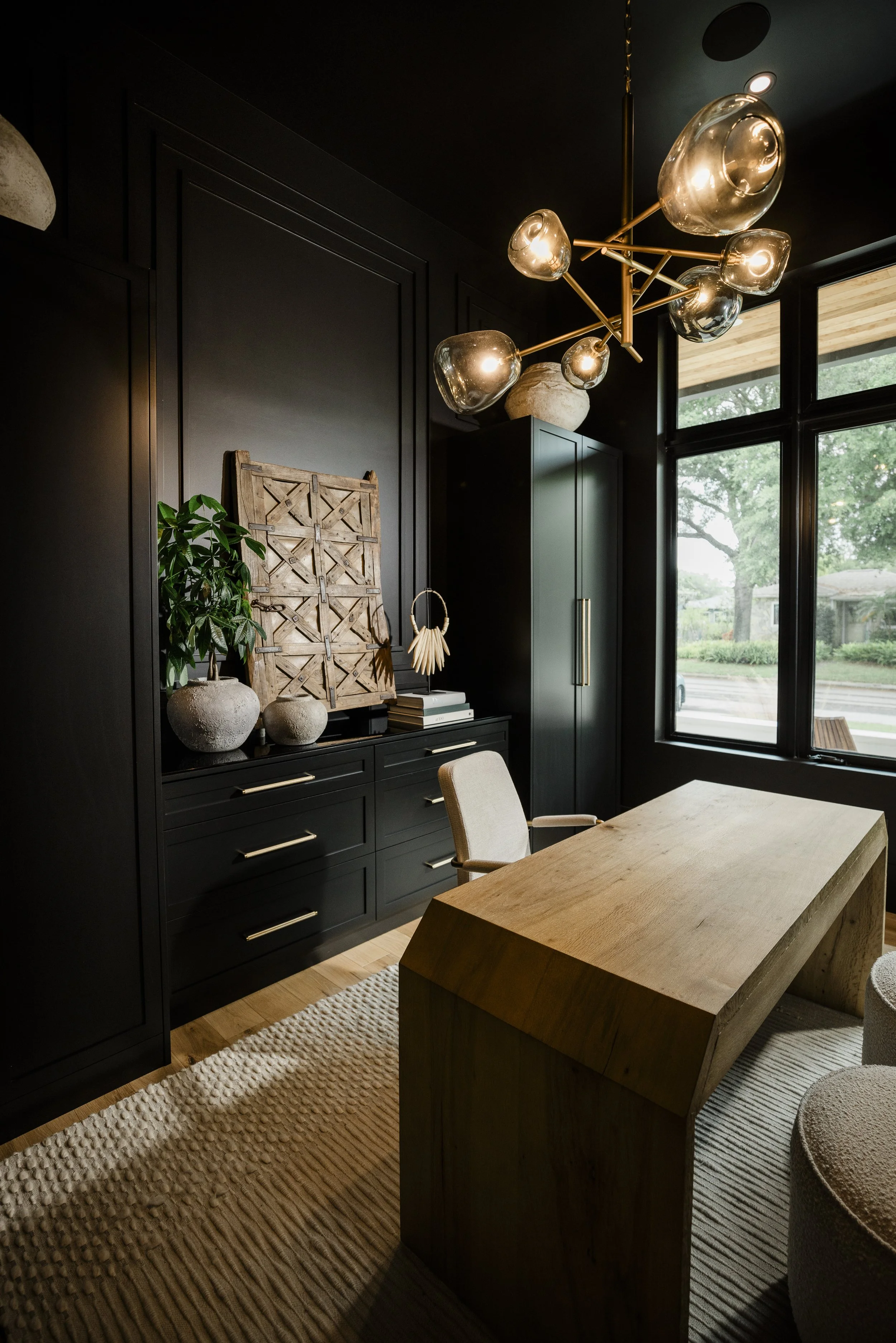 Interior of a modern dining room with a dark cabinet, decorative vases, and a wooden table. A geometric-shaped chandelier hangs from the ceiling, and large windows provide natural light and a view of greenery outside.