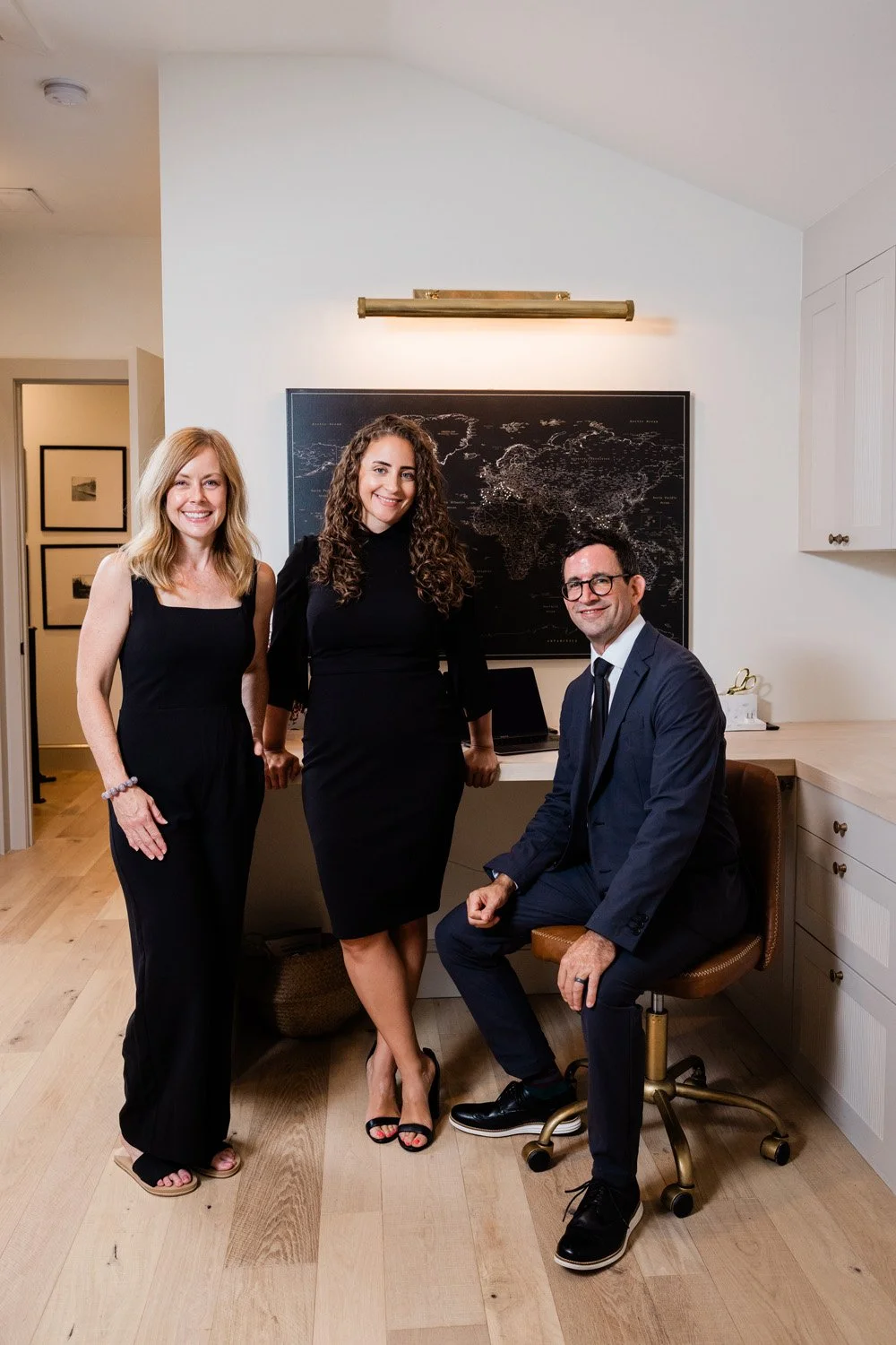 Three professionally dressed people in an office, two women standing and one man seated, smiling at the camera, a world map on the wall behind them.