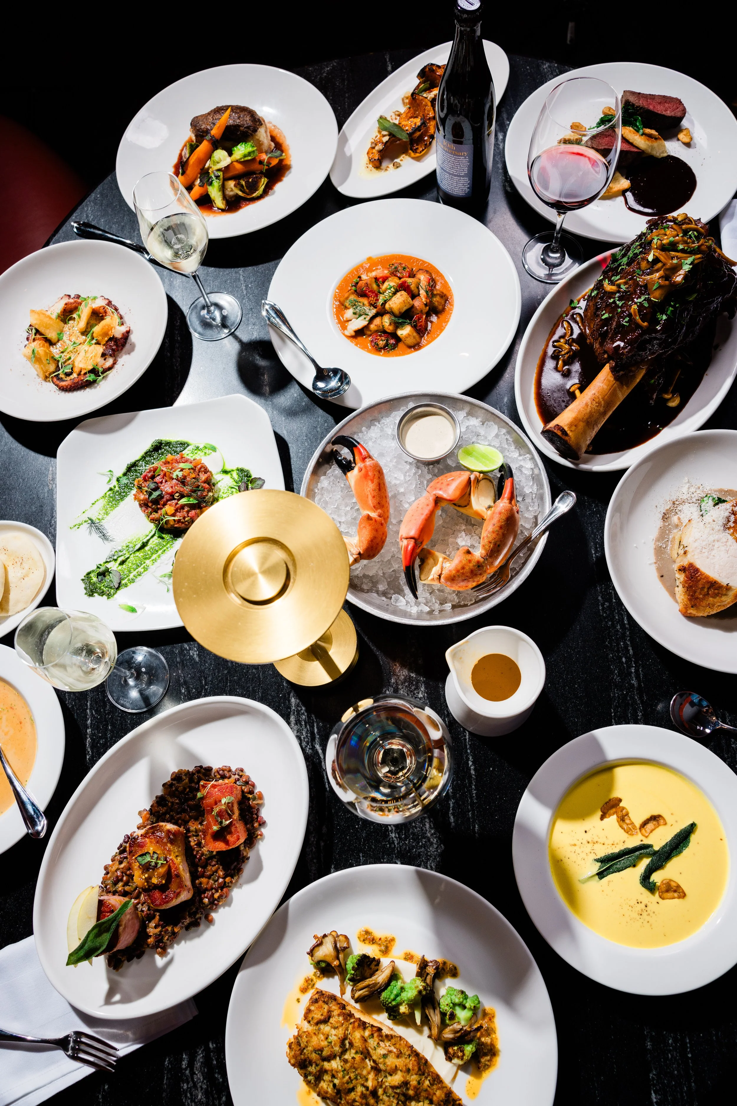 A table with various gourmet dishes, including meats, vegetables, and soups, accompanied by glasses of white and red wine, and a bottle of wine, in a fine dining setting.