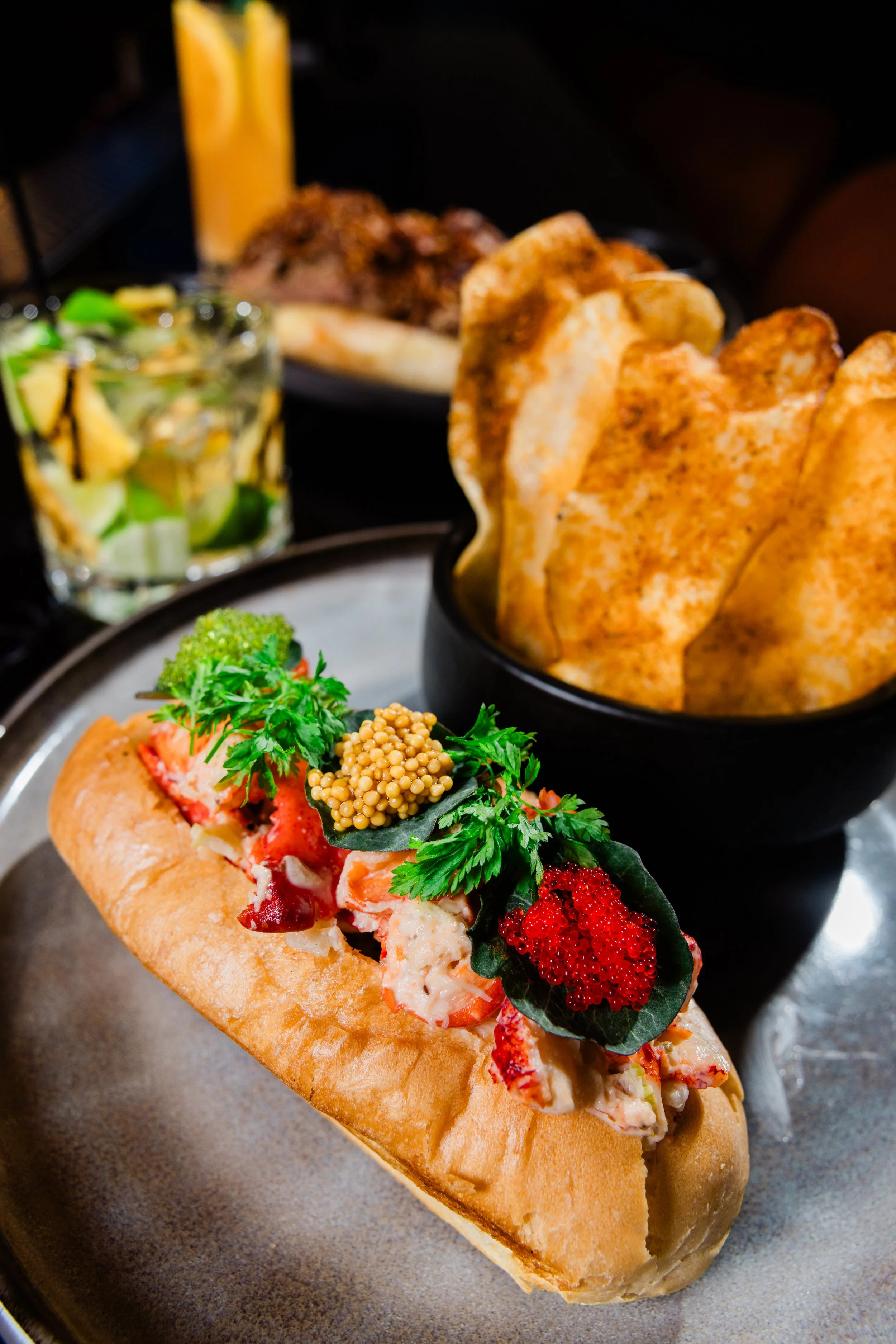 Close-up of a gourmet lobster roll with fresh herbs and caviar, served on a toasted bun, with crispy potato chips, shredded lettuce, and mustard in the background.