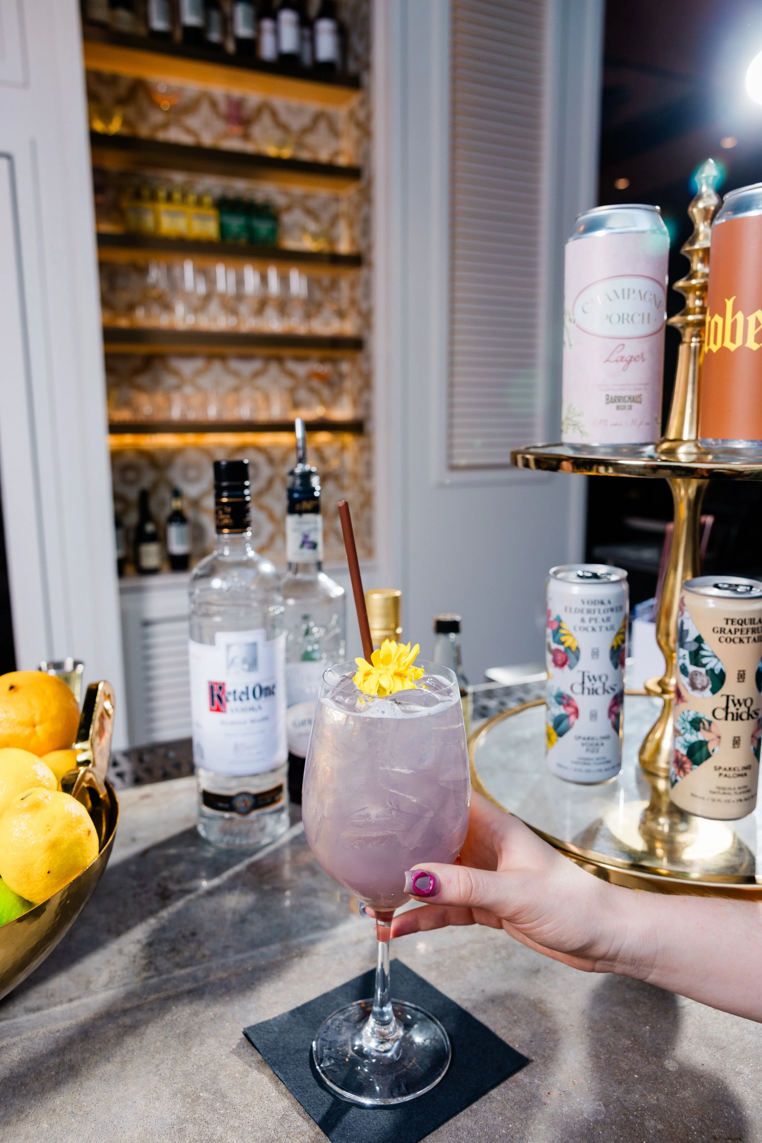 A person holding a pink cocktail garnished with a yellow flower, with a bar counter in the background containing bottles, lemons, and cans.