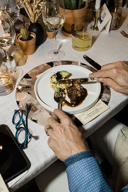 Person cutting food on a plate at a dinner table with wine glasses, small potted plants, and silverware.