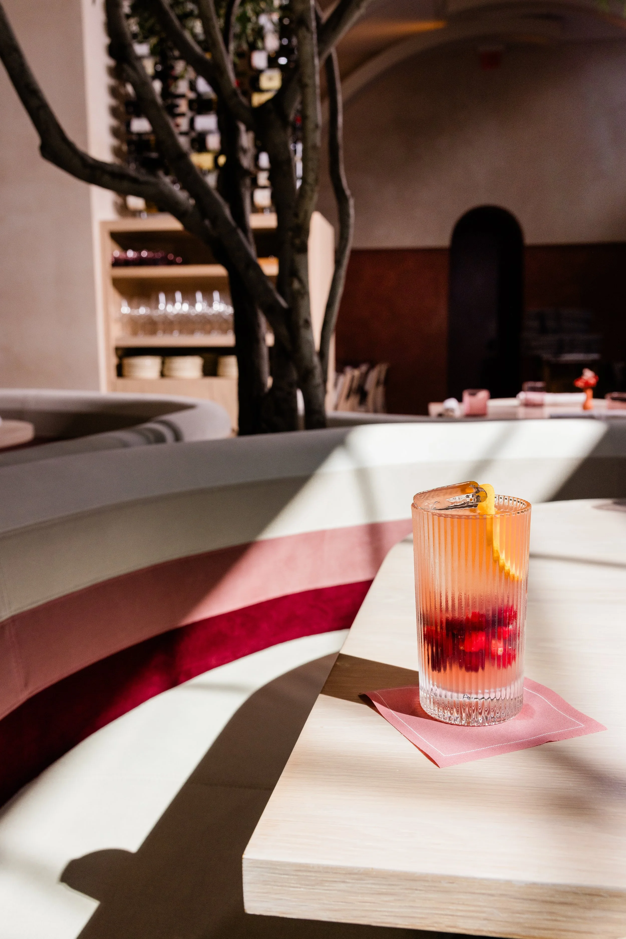 A pink and red layered drink with a slice of orange and a straw on a pink napkin on a white table in a restaurant with a wine rack and glasses in the background.