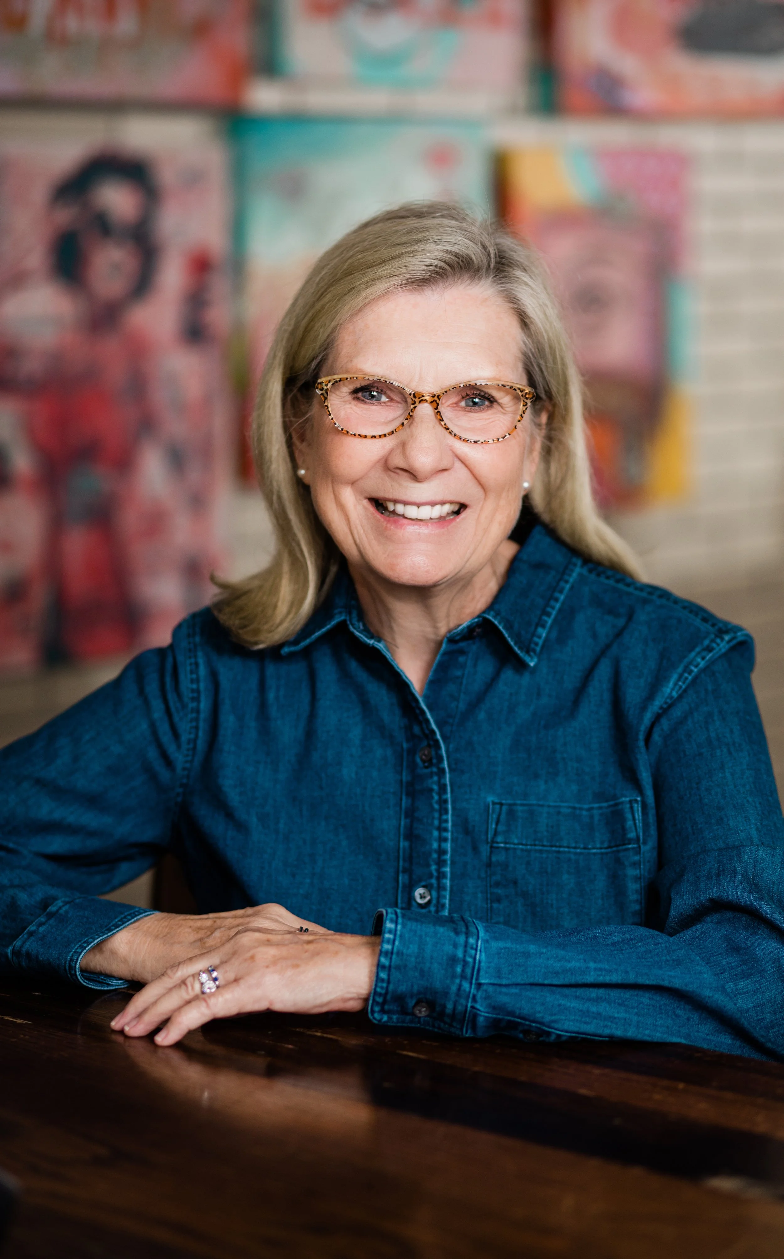 A blonde woman wearing glasses and a denim shirt poses at Elevage with contemporary art in the background