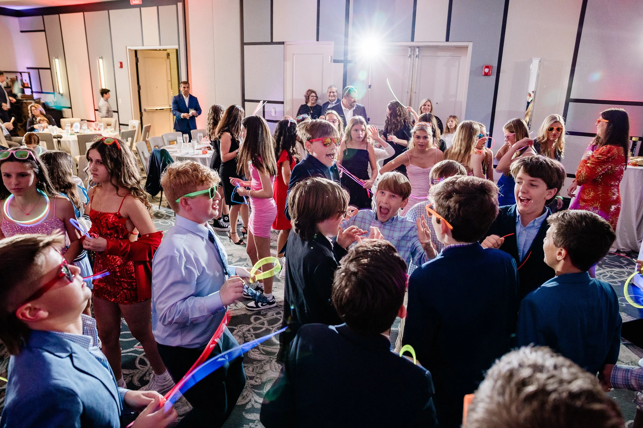 A large group of children, mostly girls, at a party or celebration, wearing colorful dresses and sunglasses, with some holding glow wands, and adults in the background in a decorated indoor venue.