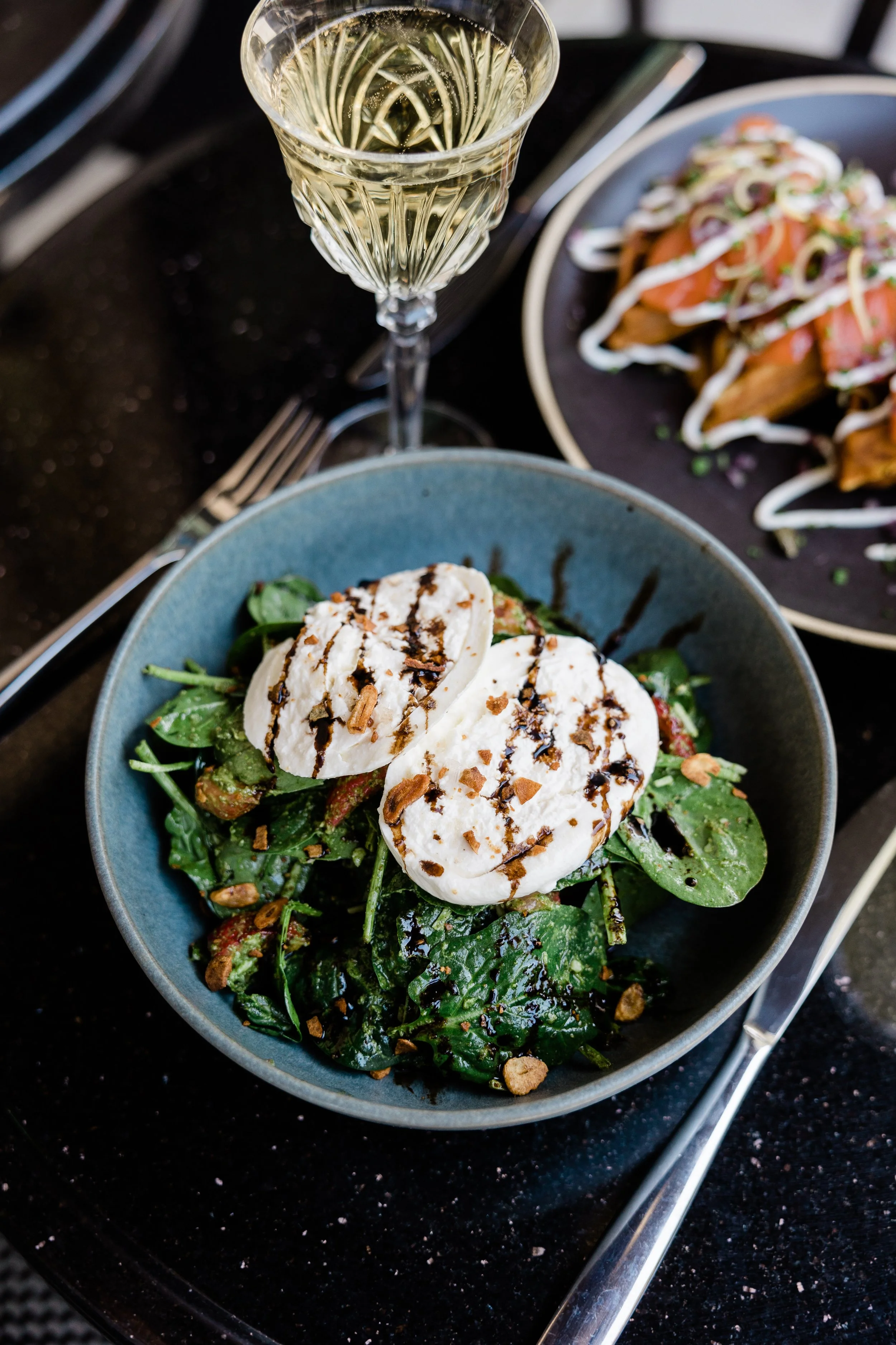 A glass of white wine, a salad with greens, cheese slices, and balsamic glaze, and a plate of what appears to be salmon topped with sauce and garnished. Silver utensils are visible on a black surface.