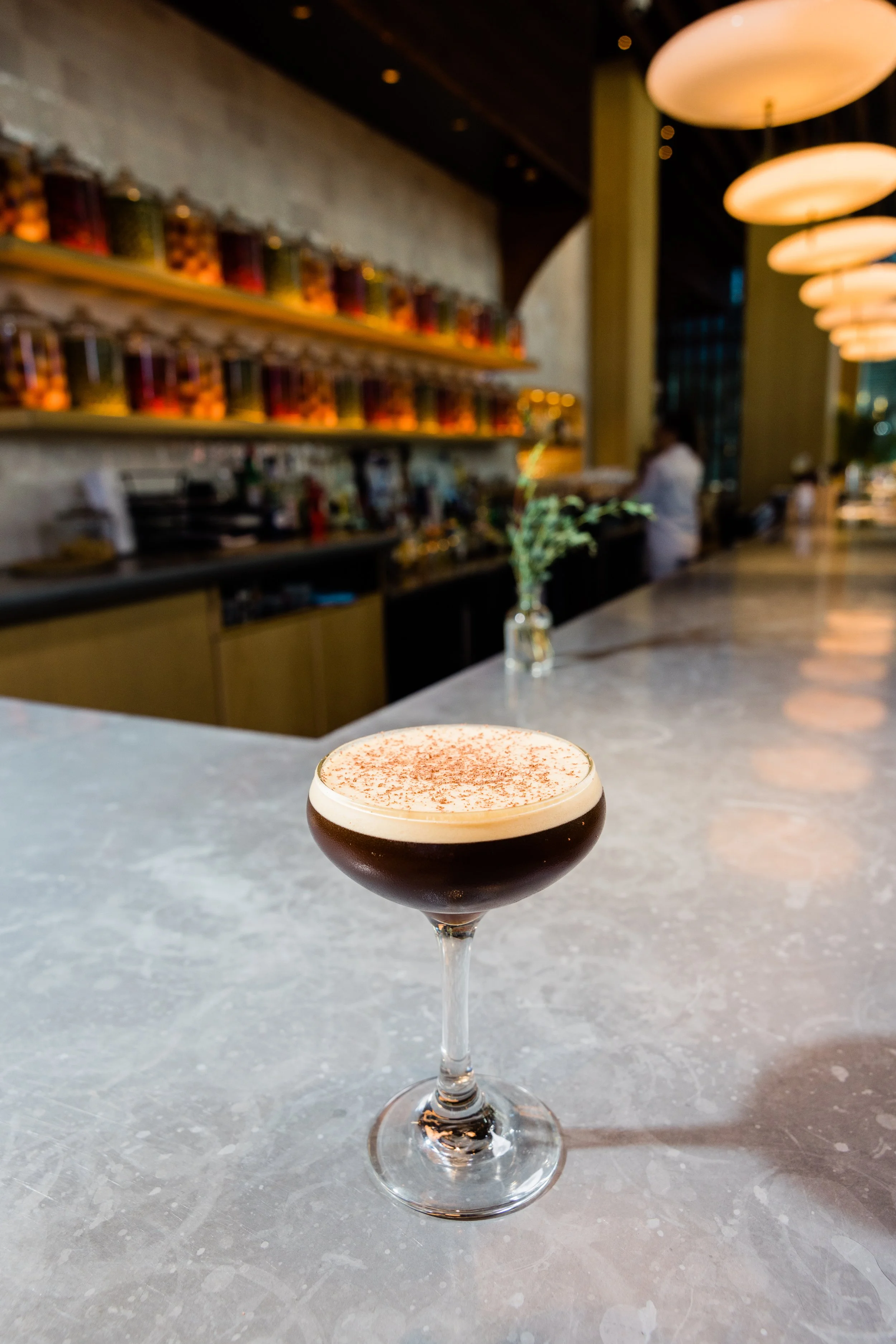 A cocktail drink in a stemmed glass with frothy cream and cocoa powder on top, on a gray marble bar counter in a modern, warmly lit restaurant or bar.