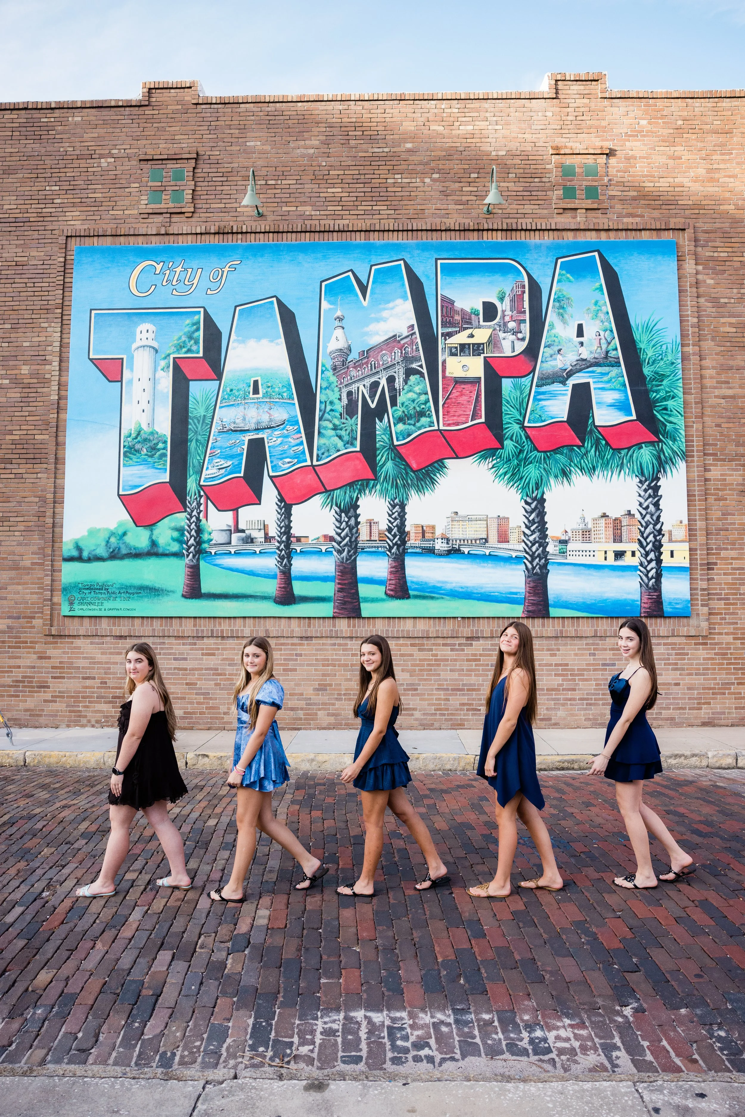 A group of five young women walking past a colorful mural on a brick building in Tampa, Florida. The mural features the word 'TAMPA' with scenes of the city, including a lighthouse, a ship, a streetcar, and people relaxing by a river, surrounded by p