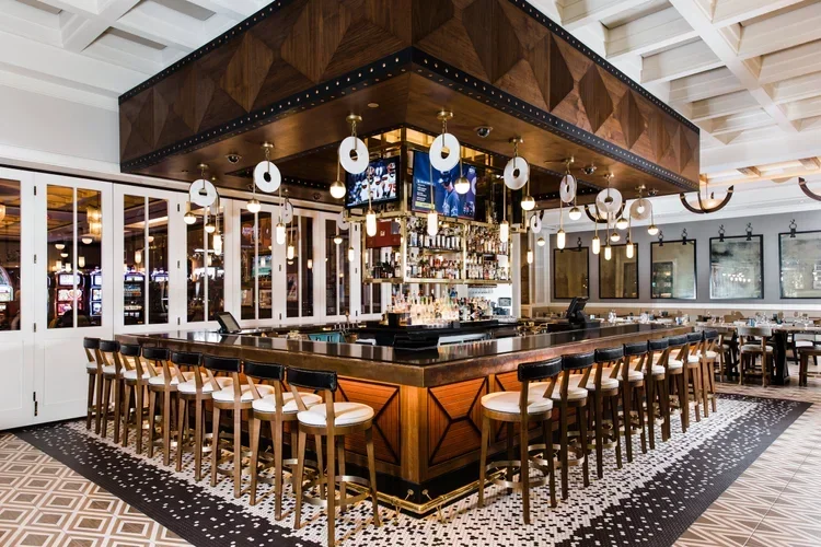 An indoor bar with a square wooden bar counter surrounded by high chairs with white seats. The bar is well-stocked with bottles, and there are hanging pendant lights above. Behind the bar are large windows, and the ceiling features a decorative grid. The floor has a mosaic tile pattern.