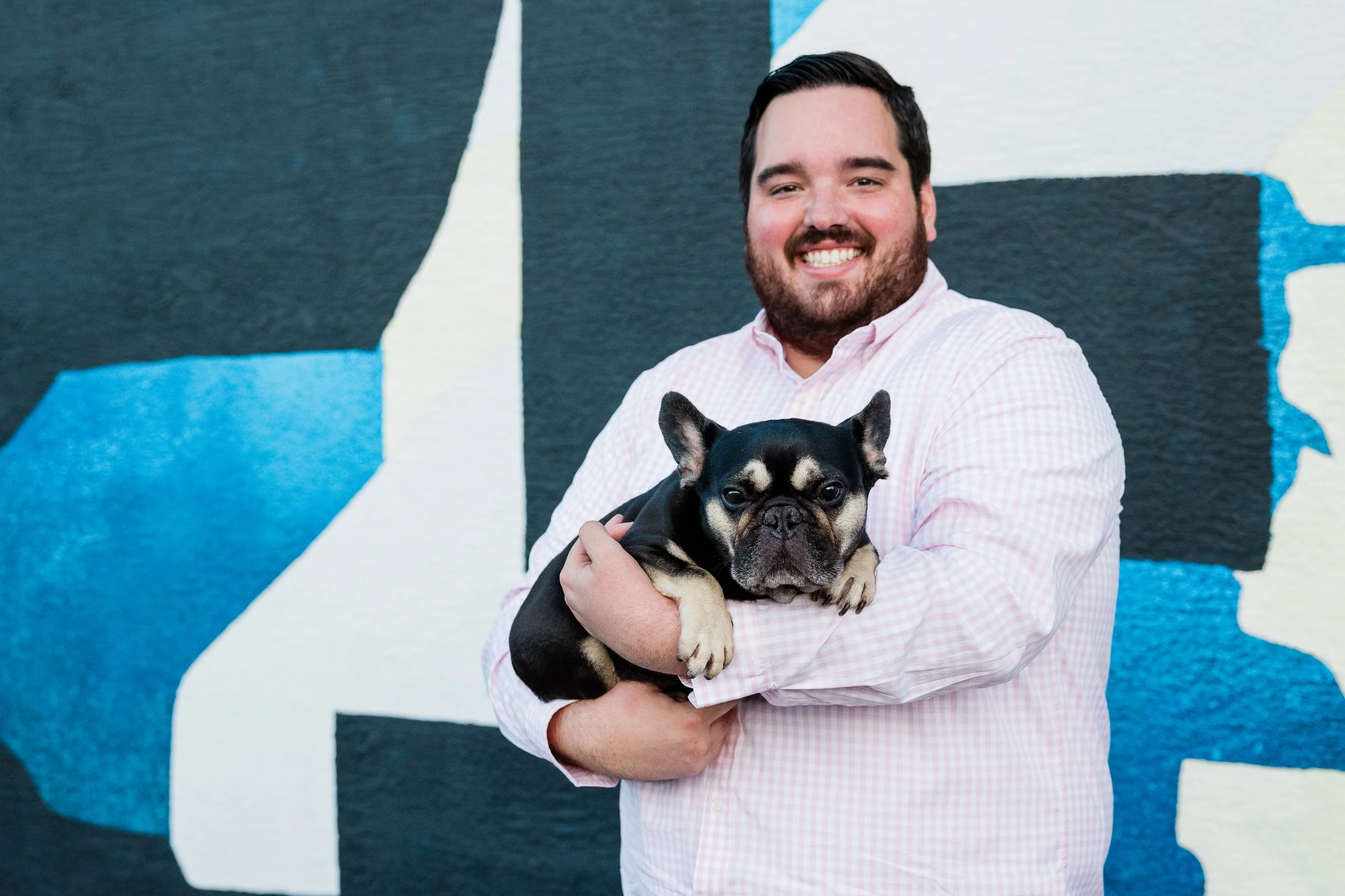 A smiling man with a beard wearing a light pink checkered shirt holding a black and tan French Bulldog puppy in front of a colorful mural with abstract shapes in blue, black, white, and beige.