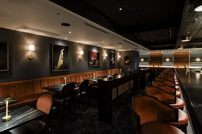 A dimly lit bar or restaurant with dark walls, framed artwork, a long orange-brown banquette, and a row of orange armchairs along the bar counter. Bern's Steak House bar.