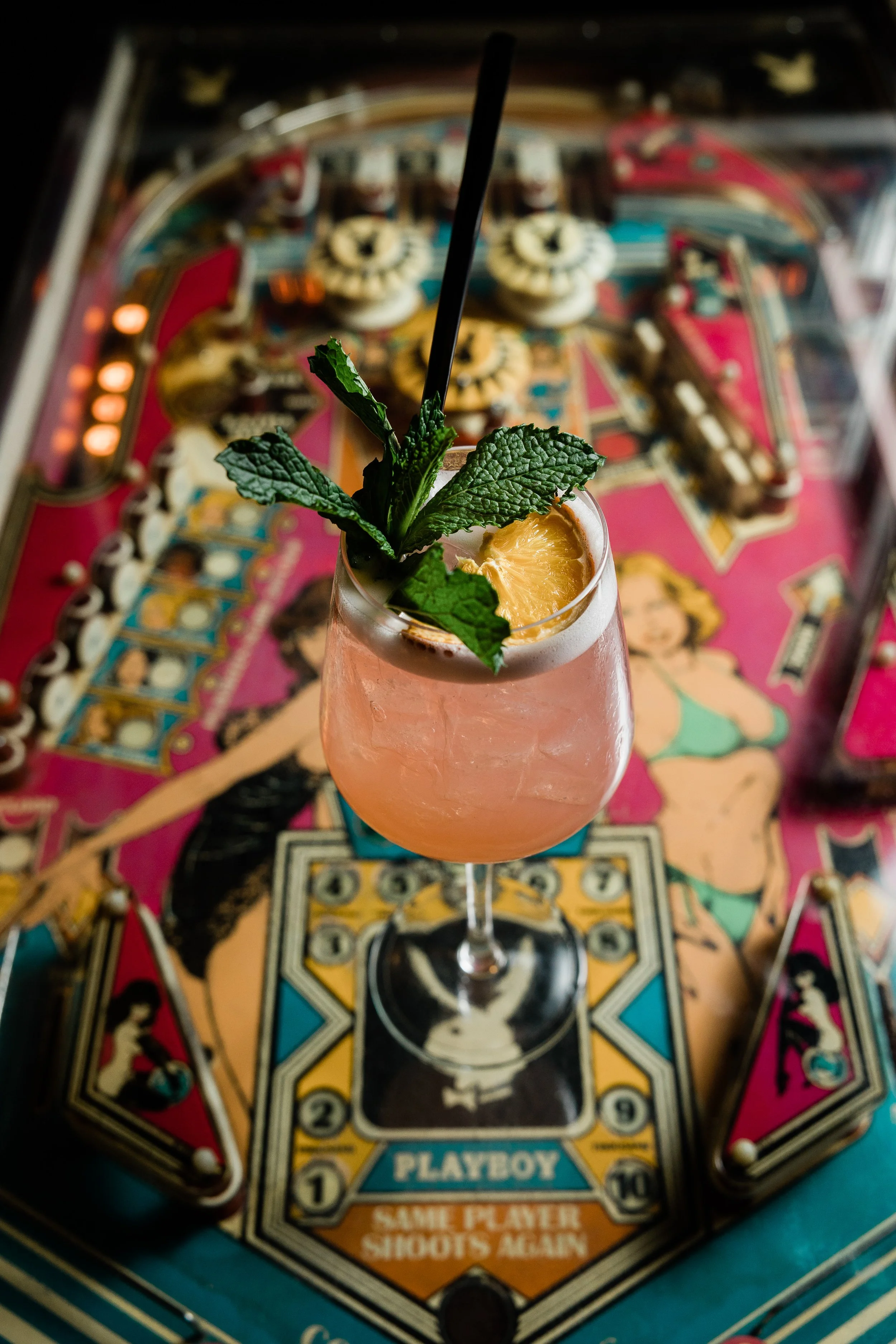 Close-up of a pink cocktail drink garnished with fresh mint leaves and a slice of pineapple, with a pinball machine game in the background.