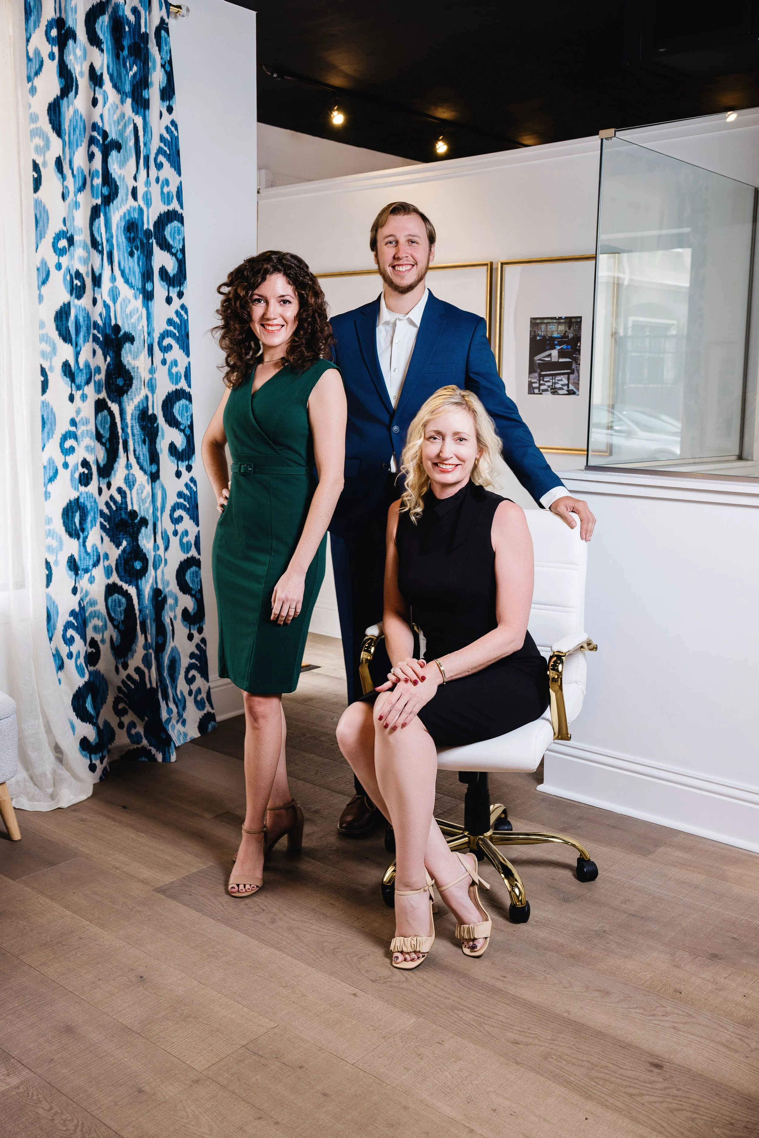 Four people in formal attire posing in an art gallery or office space, with a blue and white curtain on the left and framed artwork on the wall in the background.
