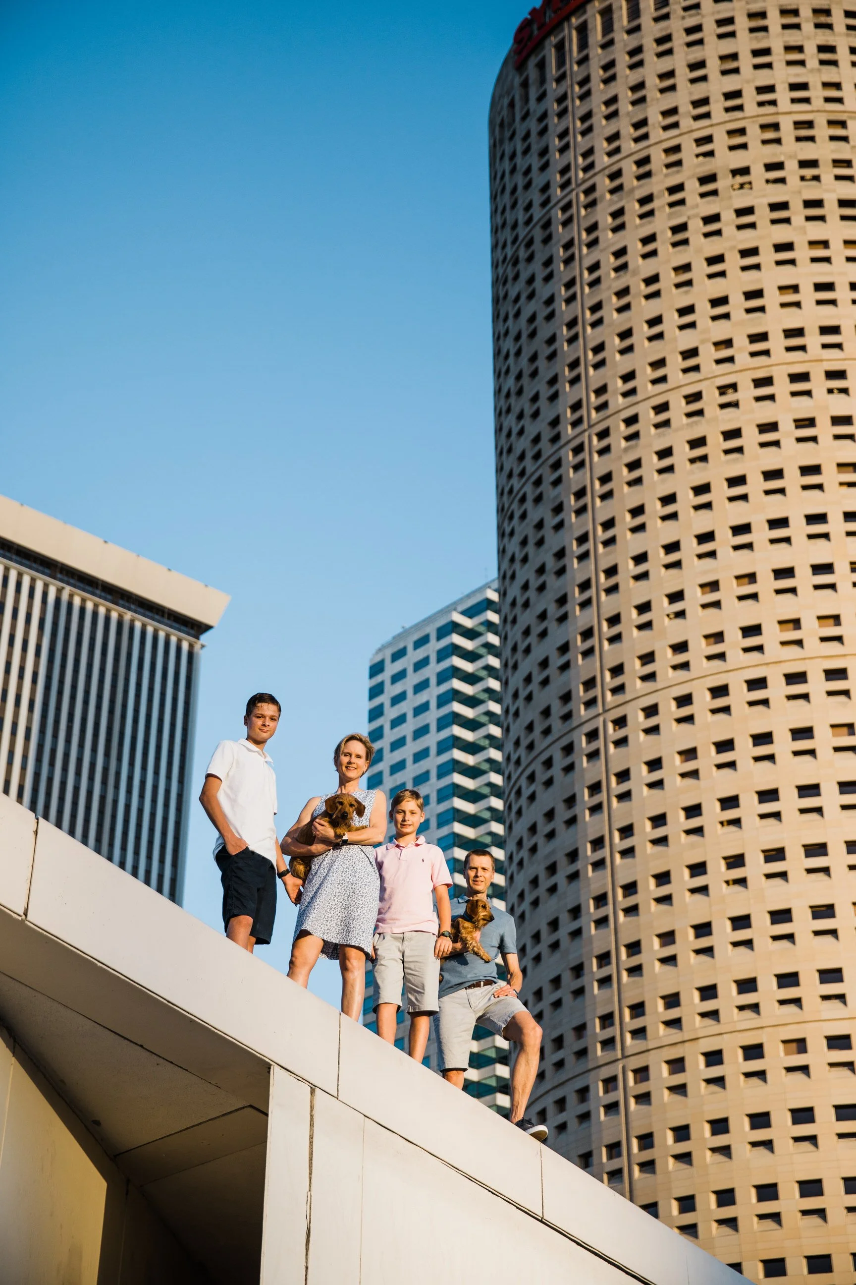 A group of five people and two dogs standing on a white balcony ledge in front of tall modern city buildings under a clear blue sky.