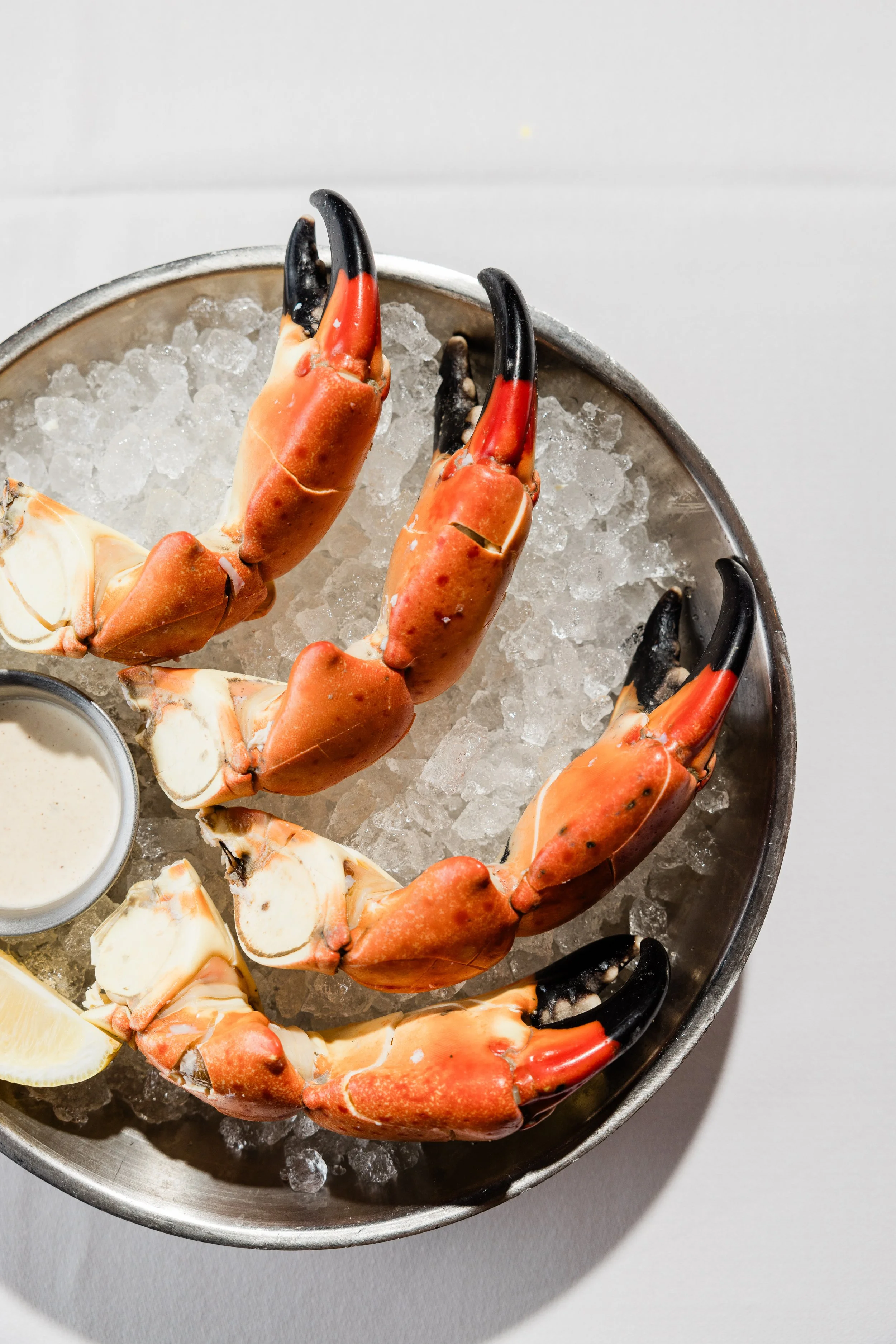 Close-up of cooked crab claws on ice in a metal dish, with lemon wedge and dipping sauce nearby.