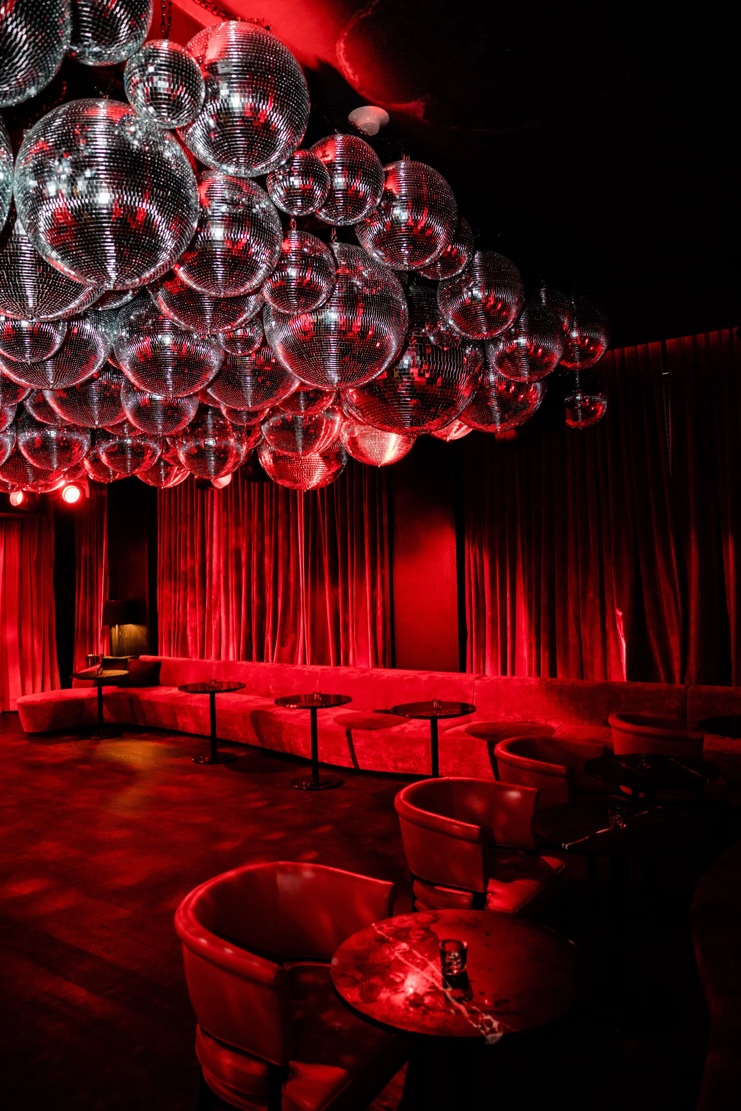 A dimly lit lounge or club with red curtains and numerous disco balls hanging from the ceiling, reflecting red light across the room. The room features a curved red velvet sofa and small round tables with chairs, set against dark flooring and a black