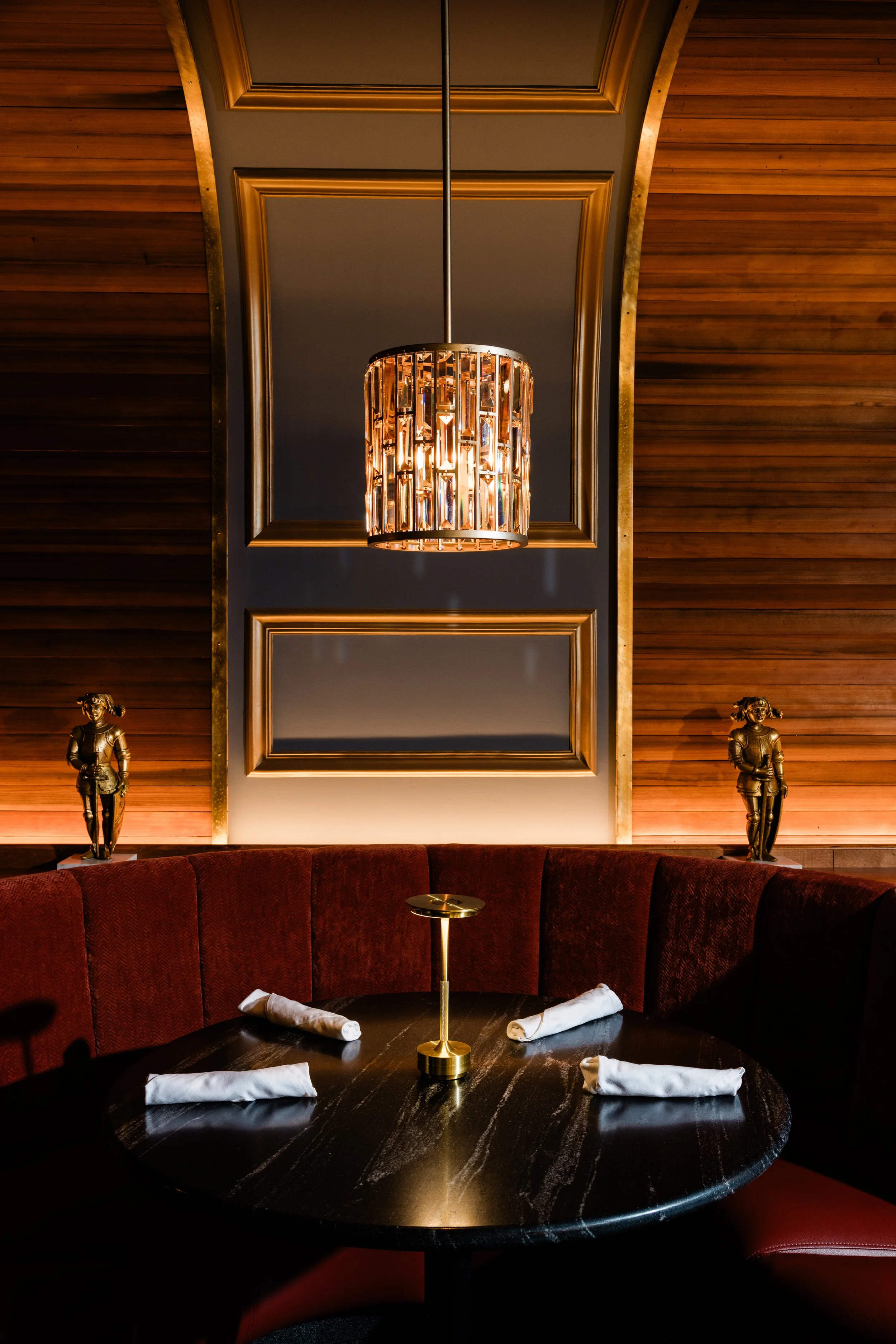 A round black marble dining table with four white napkins and a gold candle holder, surrounded by red upholstered curved seating, in a dimly lit room with wooden panel walls, a chandelier, framed mirror, and two small statues. Bern's Steak House bar.