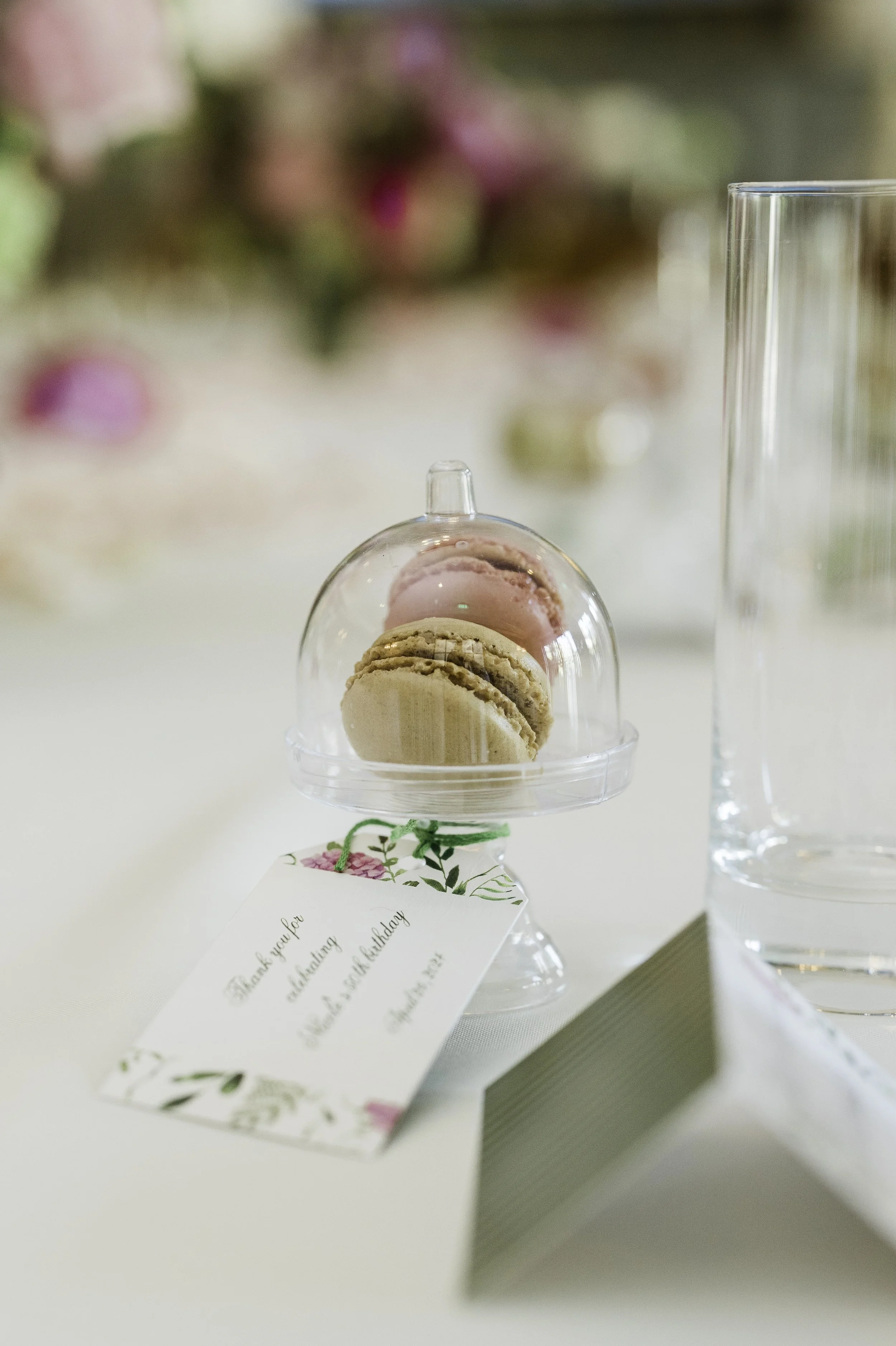 A small glass dome with macarons inside, a thank you card beside it, and a tall glass of water on the right, with a blurred floral background.