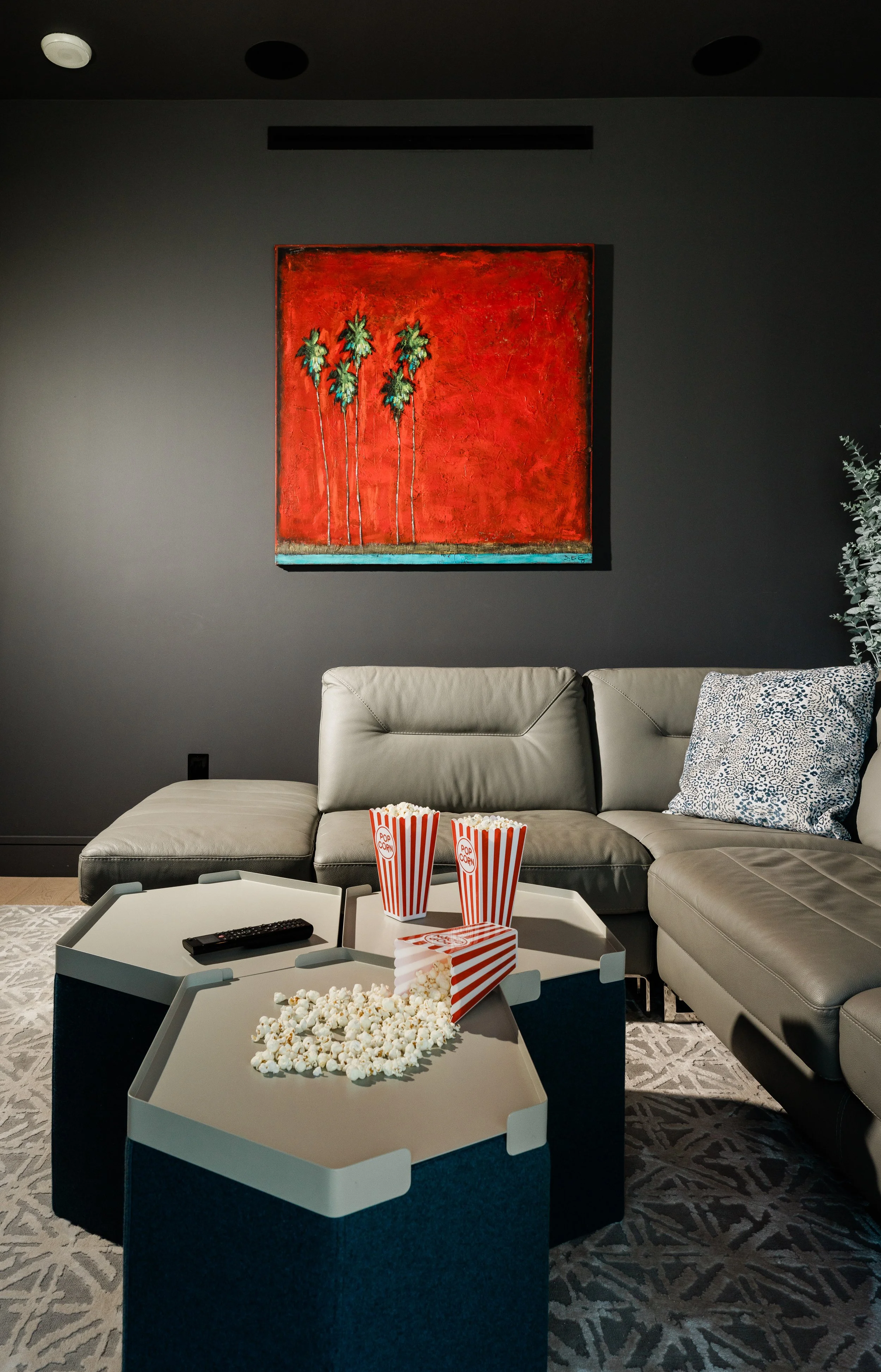 Living room with a gray sectional sofa, patterned pillow, abstract painting with palm trees on dark gray wall, and popcorn on coffee tables.