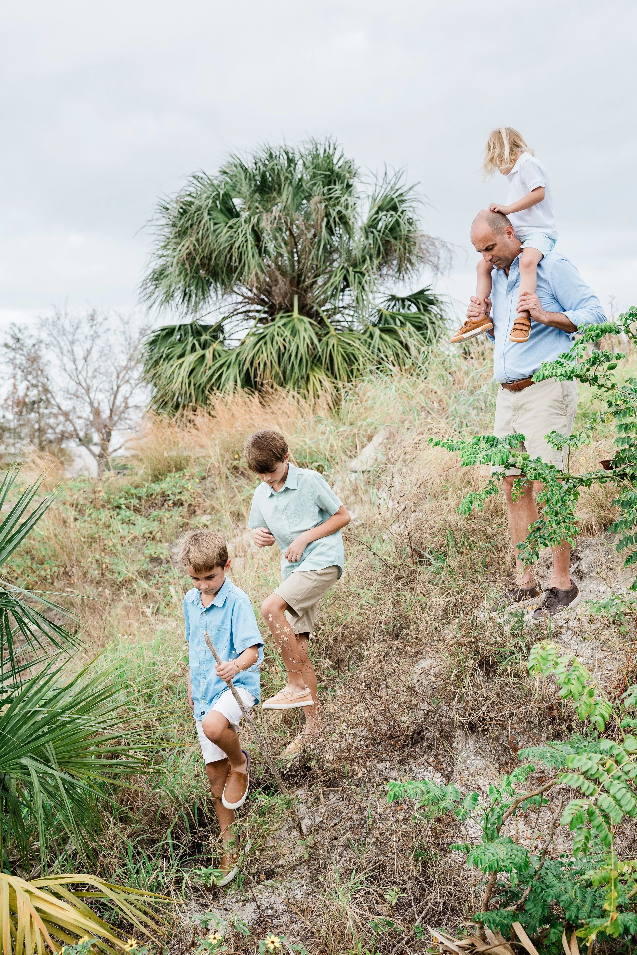 A man carrying a young girl on his shoulders and two boys walking on a hiking trail through a natural landscape with tall grasses and trees.