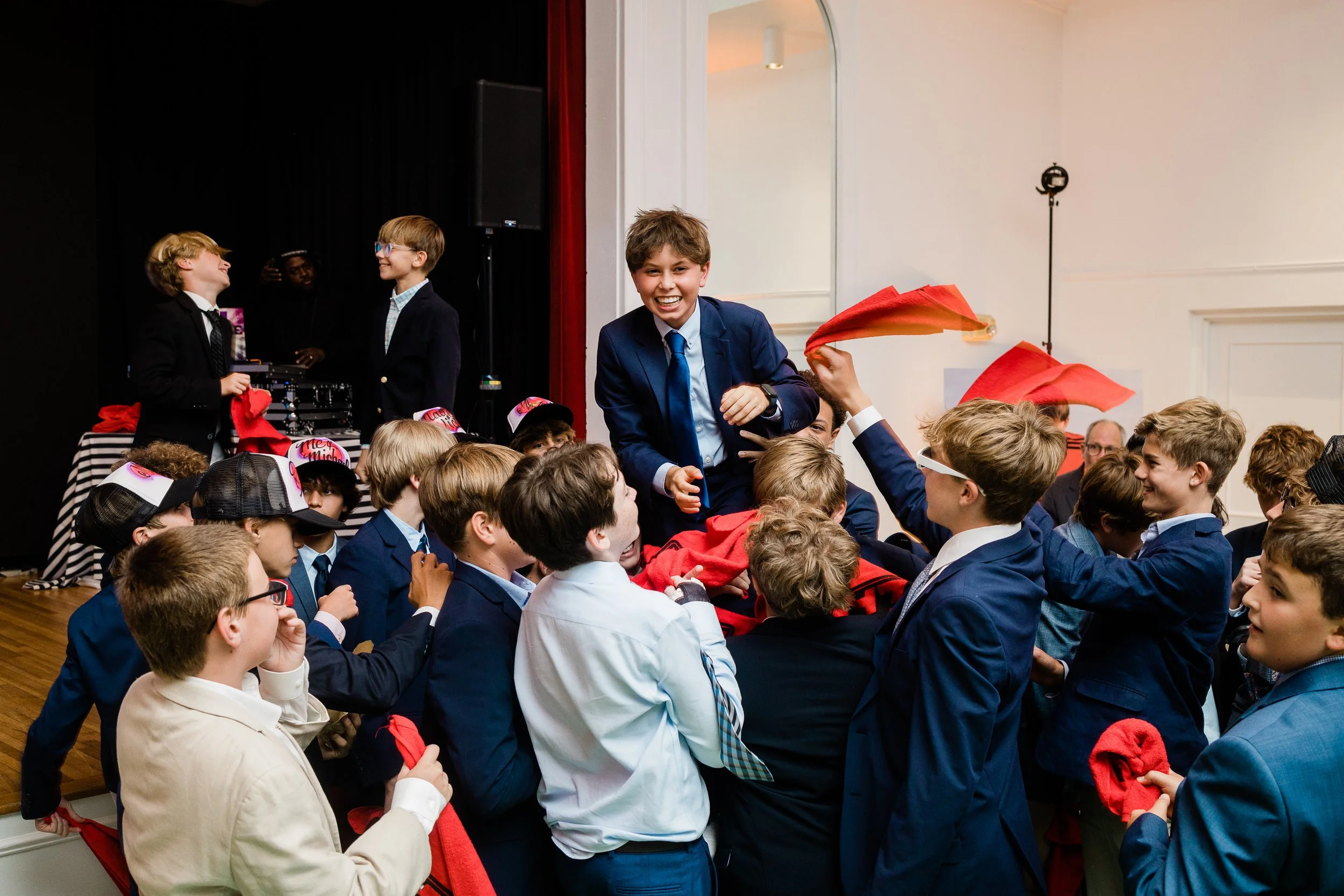 A group of children in school uniforms gathered in a room, with some wearing hats and holding red cloths. One boy is sitting on an adult's shoulder, smiling and wearing a blue jacket and tie. Other children are smiling and engaging with each other, s
