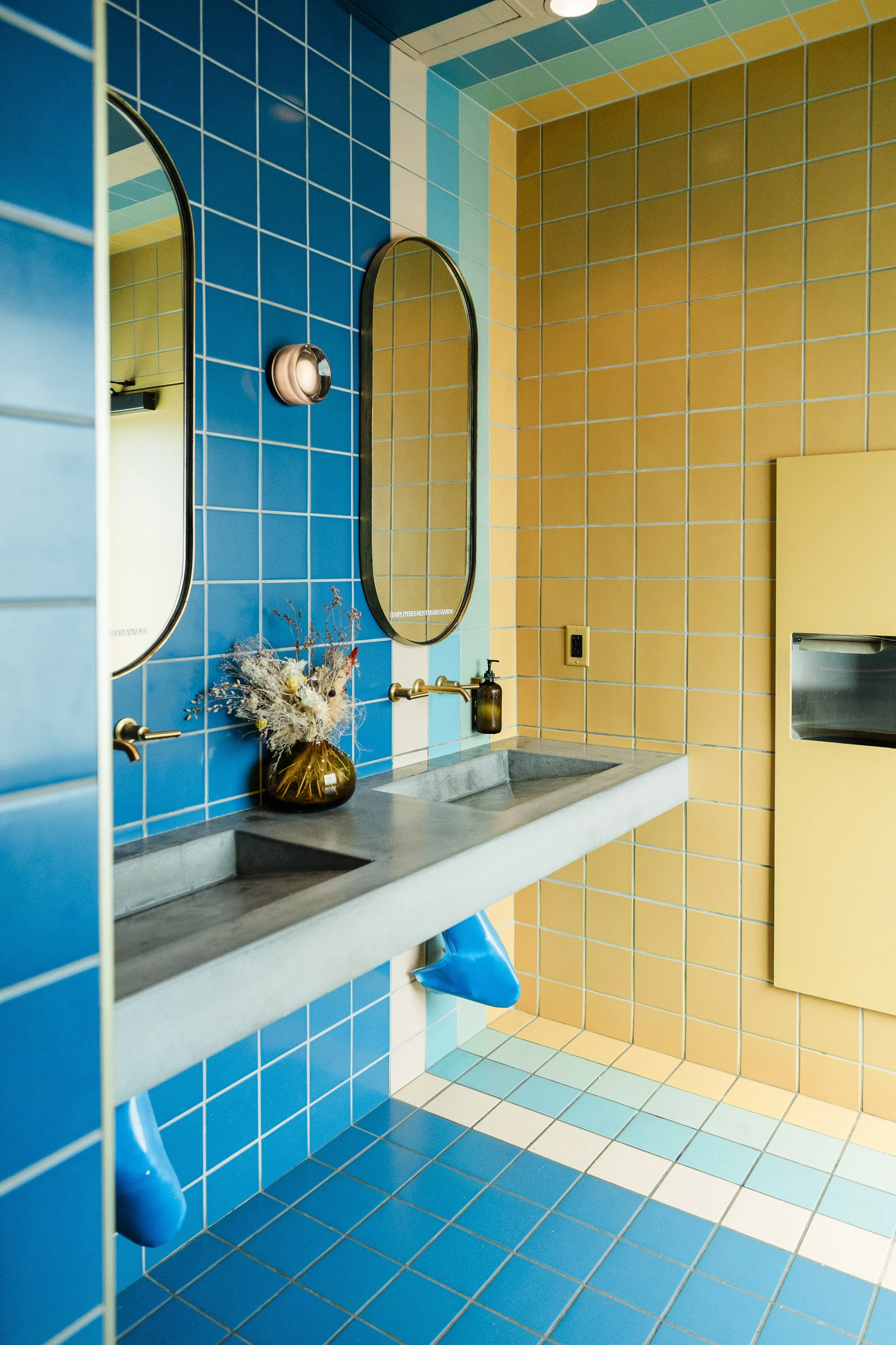Colorful tiled bathroom with blue and yellow tiles, oval mirrors, a concrete sink, a brown soap dispenser, dried flowers in a glass vase, and blue trash bags visible below the sink. Walls and ceiling are tiled in contrasting colors.