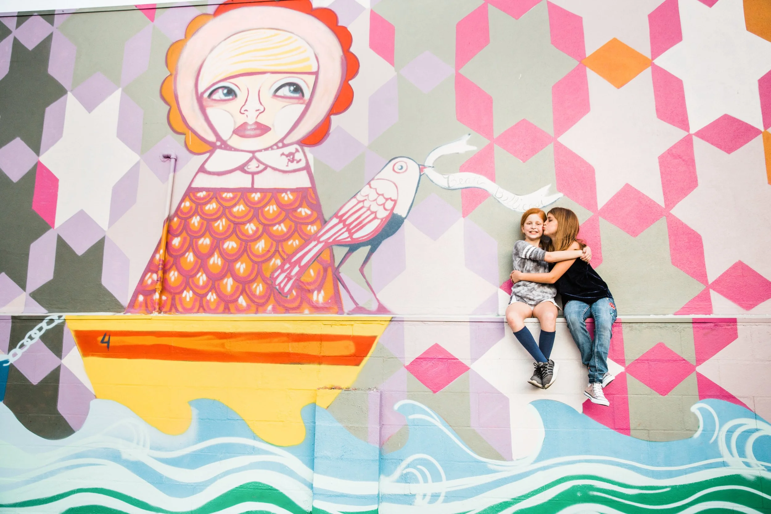 Two children, a girl with red hair and a girl with brown hair, hugging and sitting on a ledge in front of a colorful mural of a cartoon girl with a bird in her hand.
