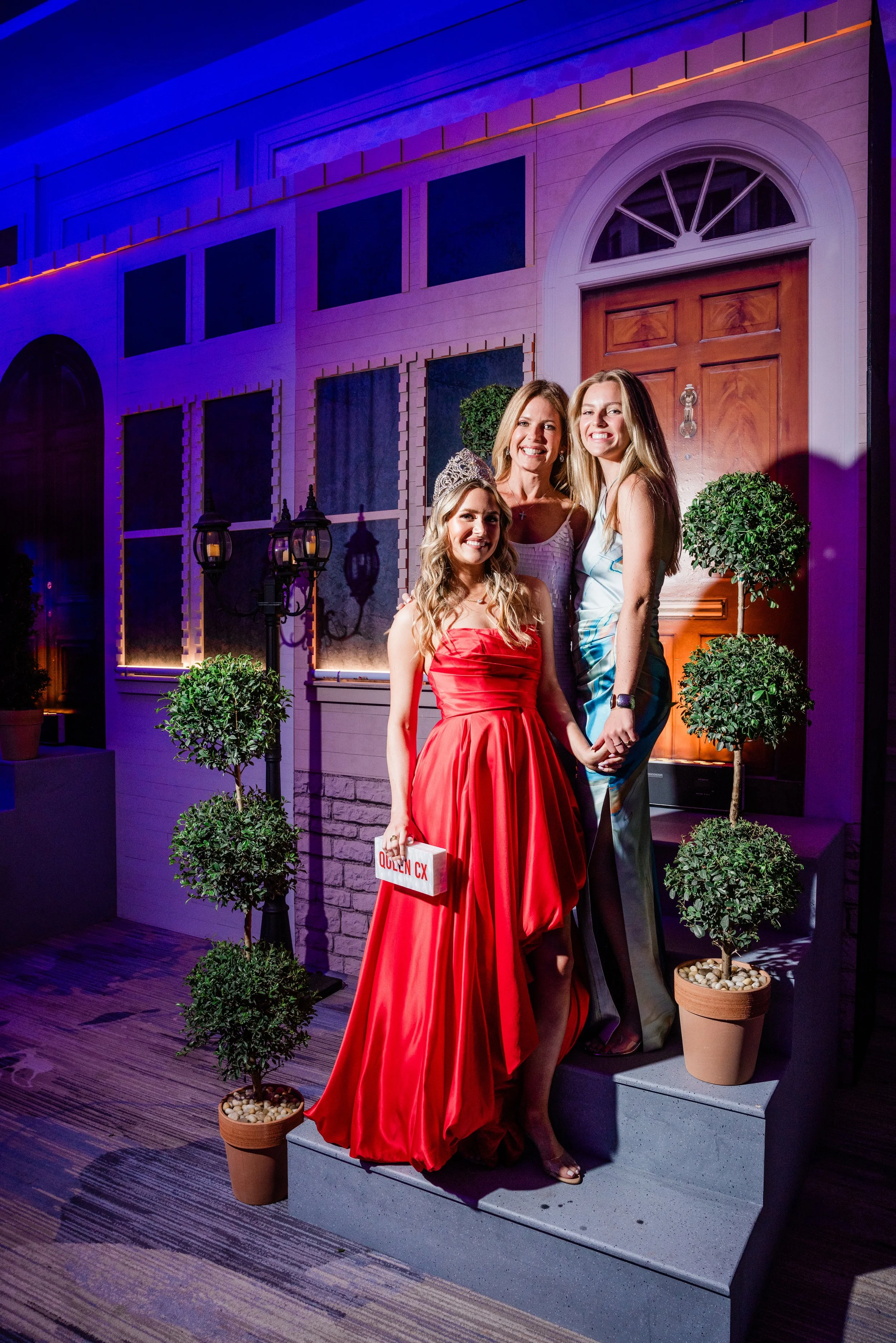 Three women standing on a step in front of a house entrance, illuminated with purple and orange lighting. One woman is wearing a red dress and a tiara, holding a sign that says "Queen CX". The other two women are smiling, one wearing a white dress an