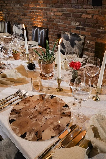 A elegantly set dining table with a marble-patterned plate, wine glasses, candles, and flower centerpieces, against a brick wall backdrop.