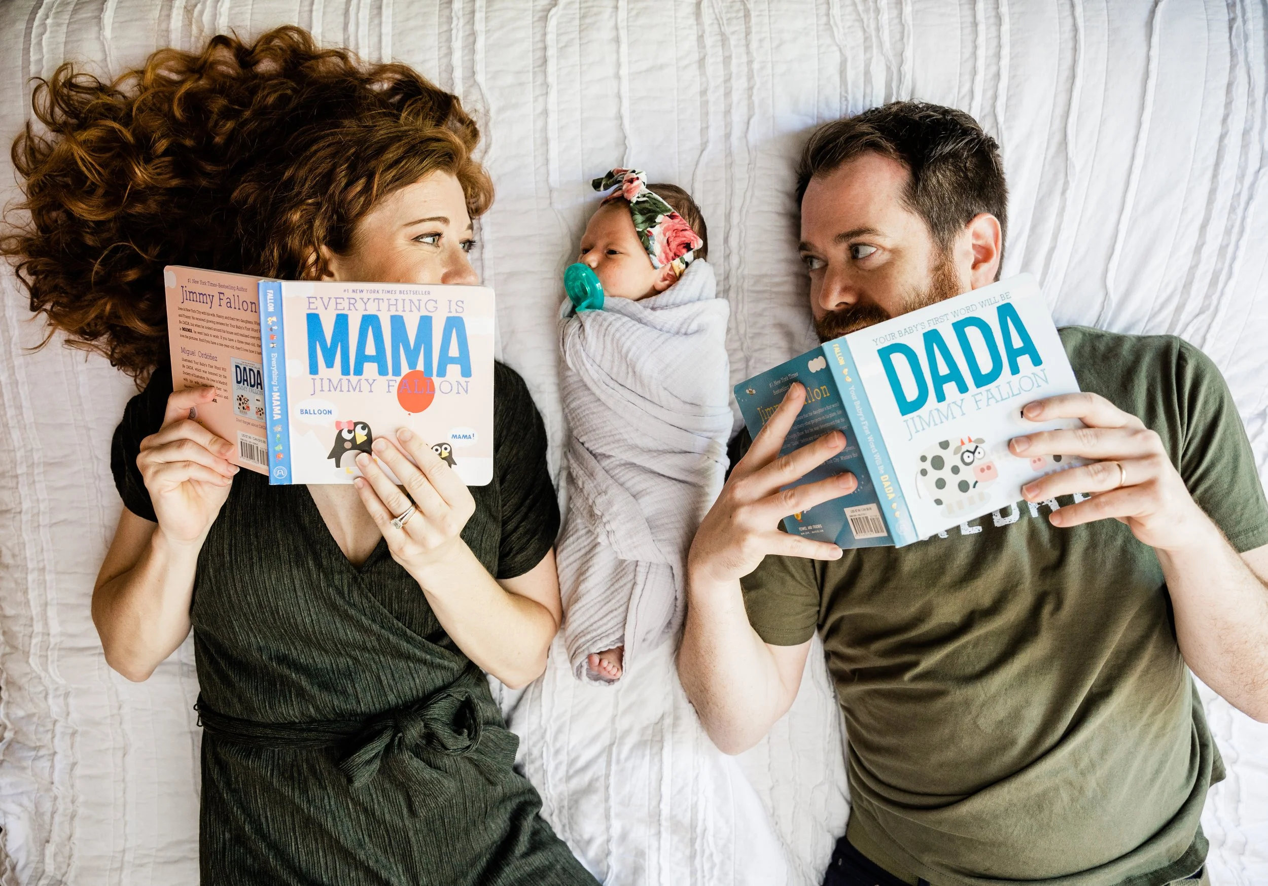New parents lie on the bed with newborn baby between them. They are readying MAMA and DADA books.