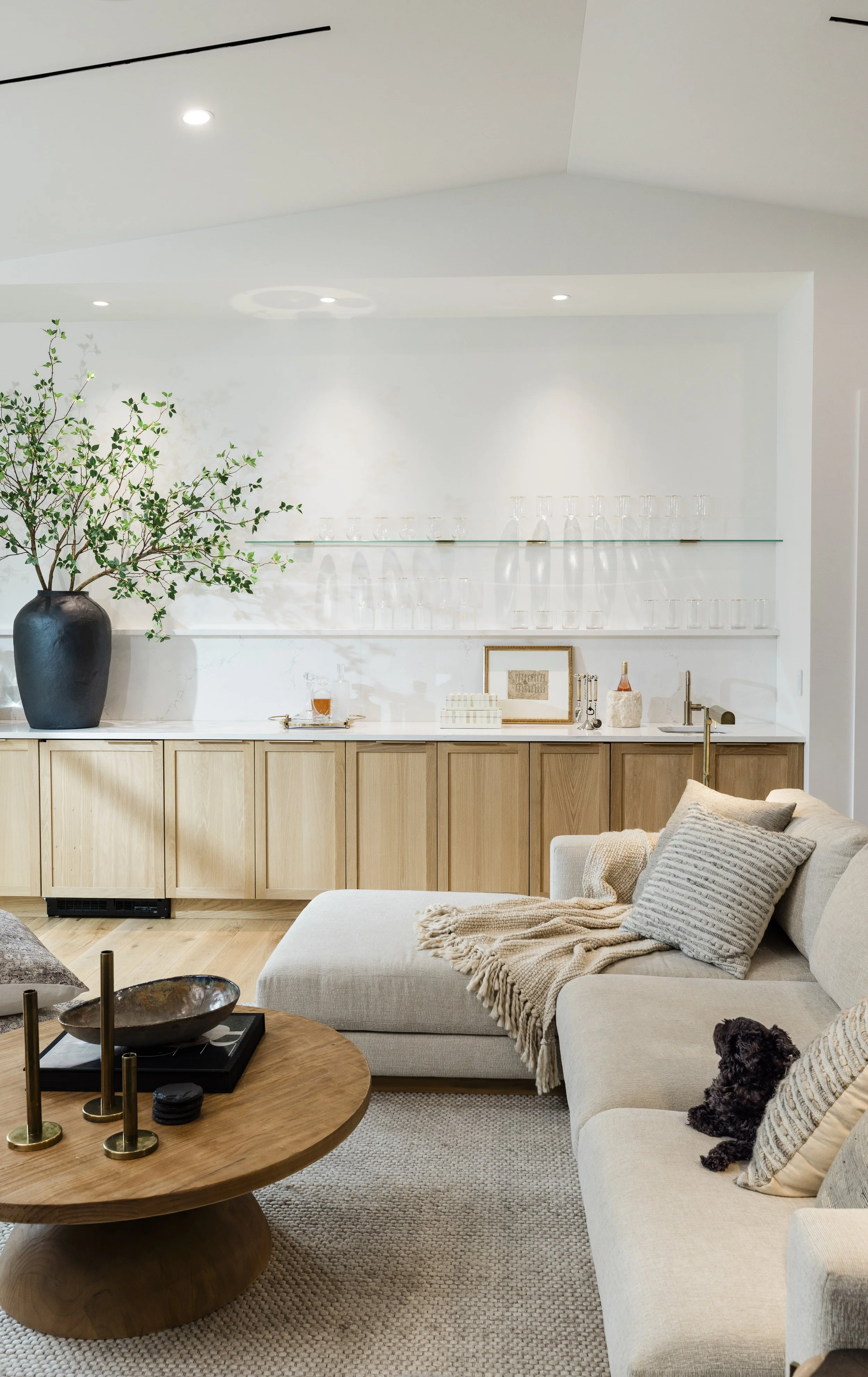 Modern living room with a beige sectional sofa, decorative pillows, beige throw blanket, wooden coffee table with accessories, and a large black vase with green leafy branches on a white countertop with glassware and framed artwork in the background.
