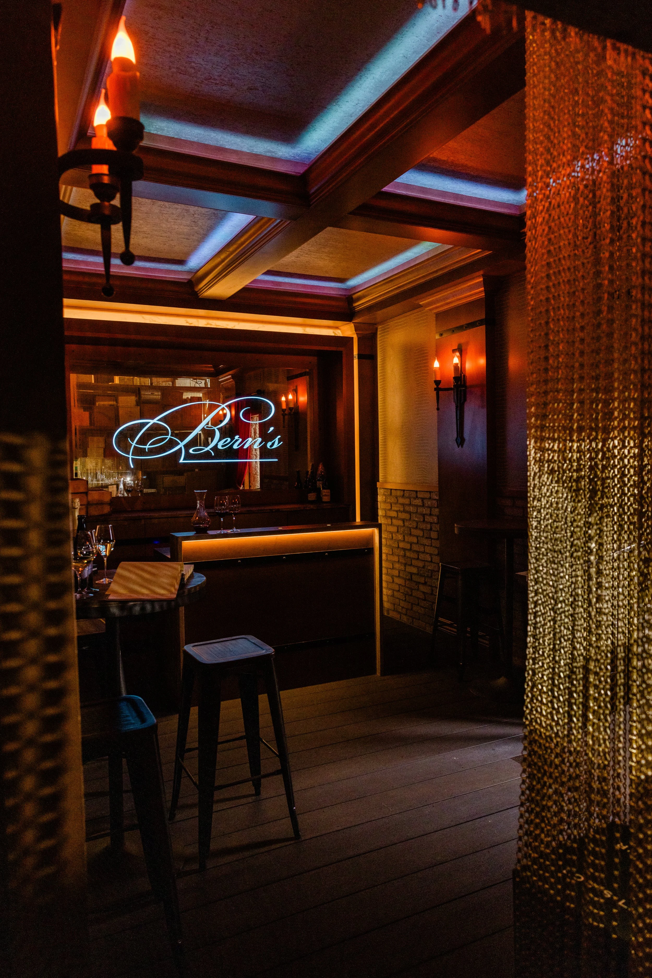 Dimly lit restaurant or bar with tables, chairs, and a decorative sign that reads "Lerni's" in neon lights. Warm lighting with wall sconces on the walls, and a textured golden curtain or wall feature.