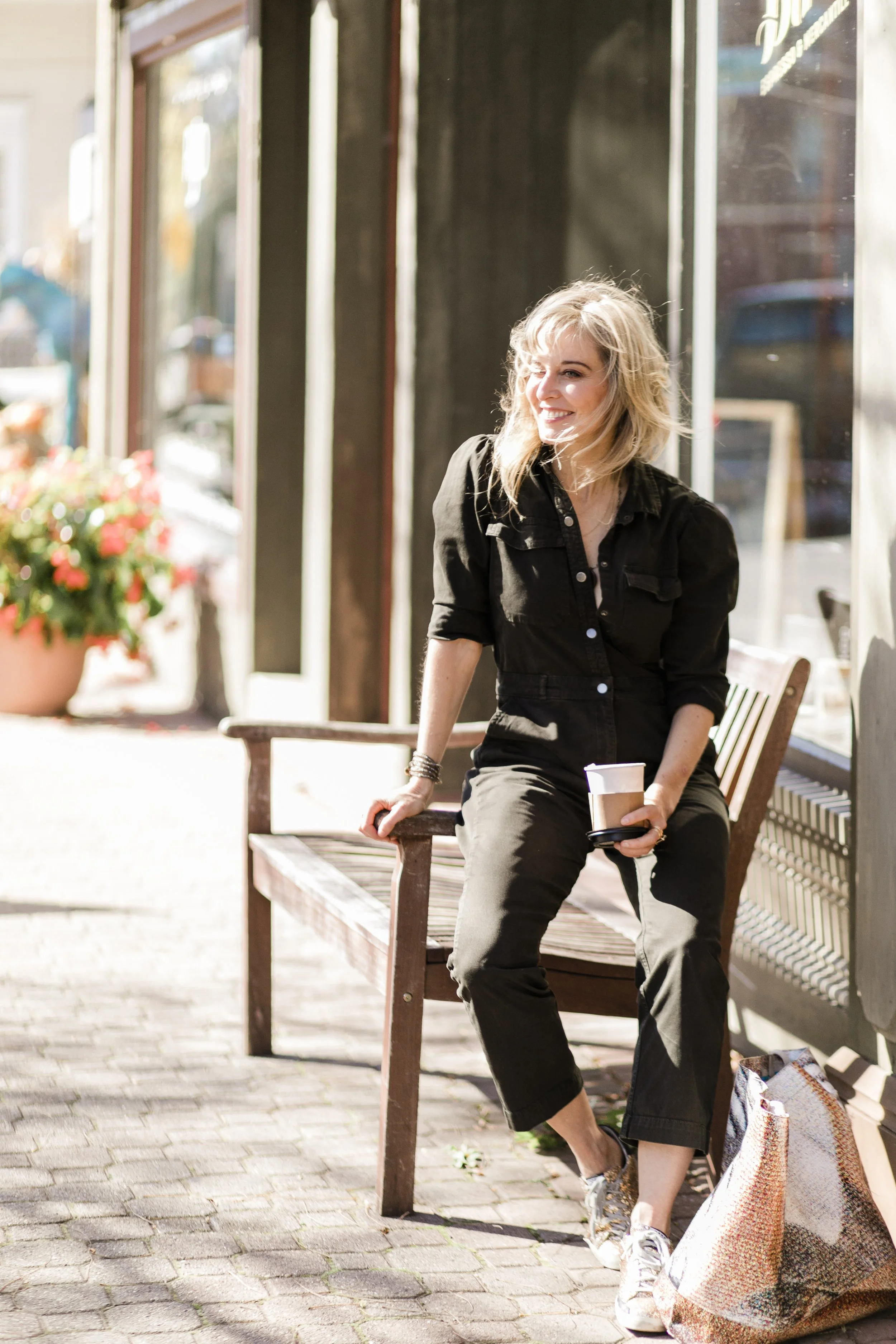 blonde woman in black jumpsuit sitting on arm of bench holding a coffee in New Jersey during a personal branding session