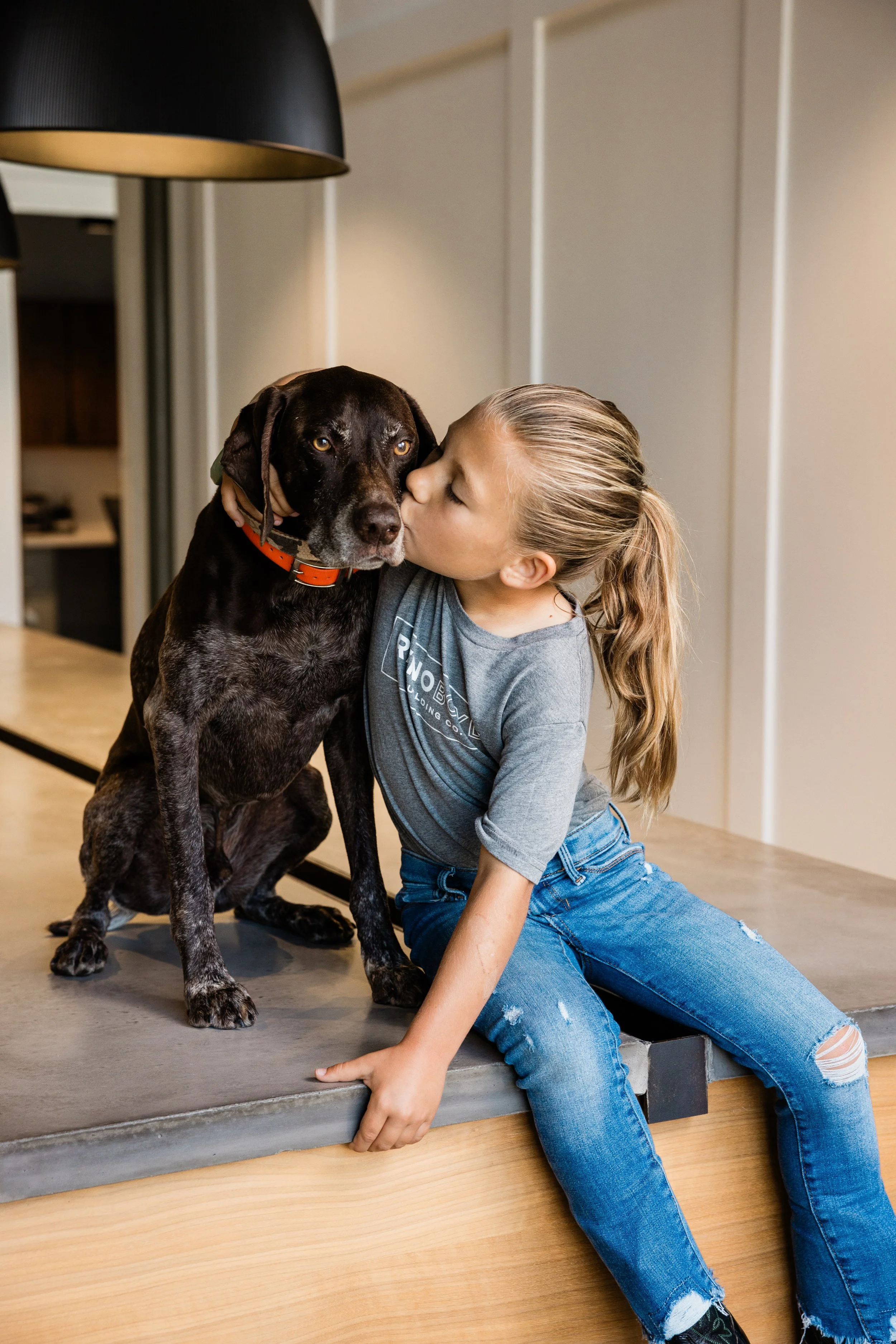 A young girl with long blonde hair and ripped jeans kneels on the floor, giving a kiss to a large black and white dog with yellow eyes, sitting on a gray platform inside a home.
