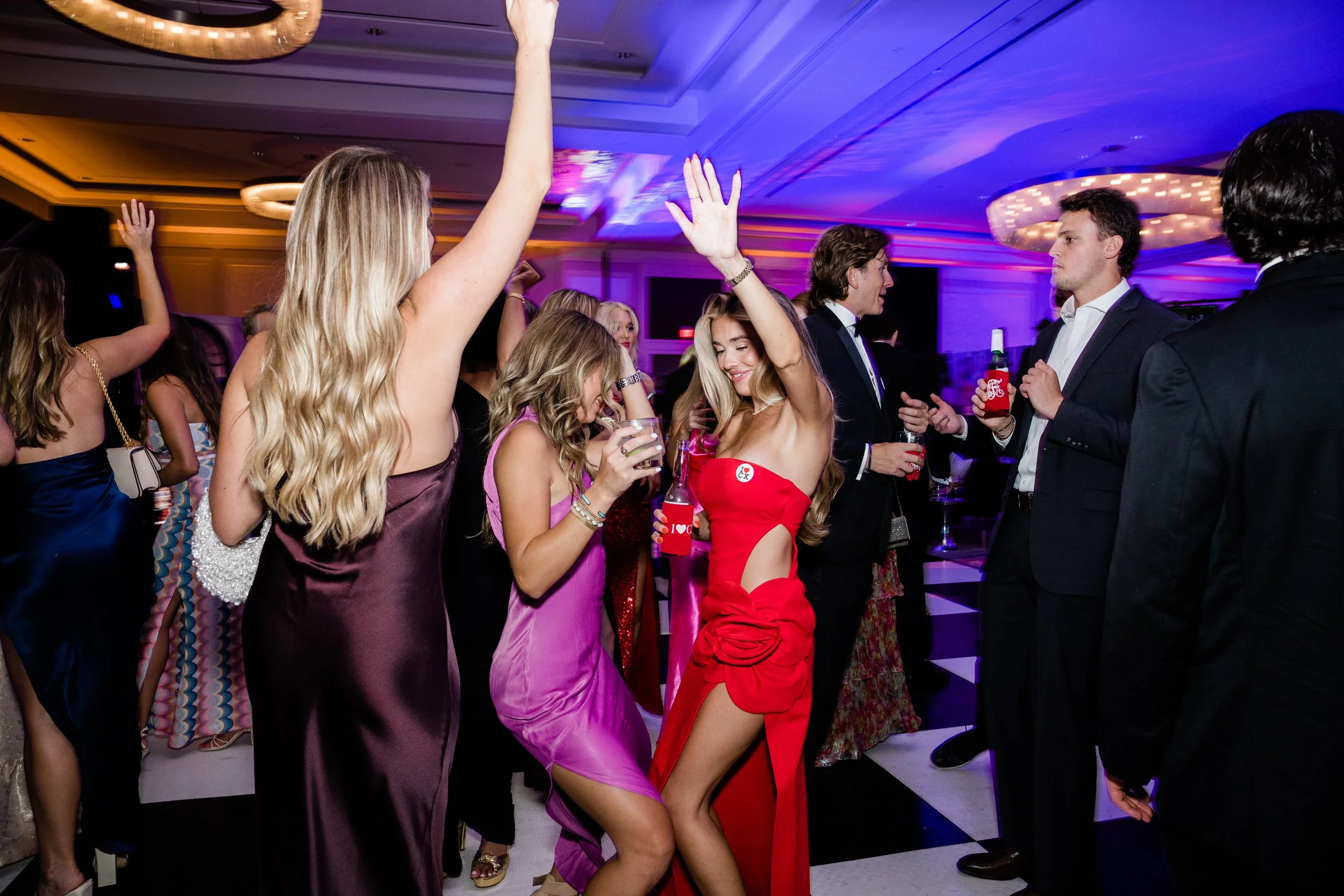 People dancing and socializing at a party or celebration inside a decorated venue with purple and pink lighting. Women are wearing elegant dresses, and men are dressed in suits.