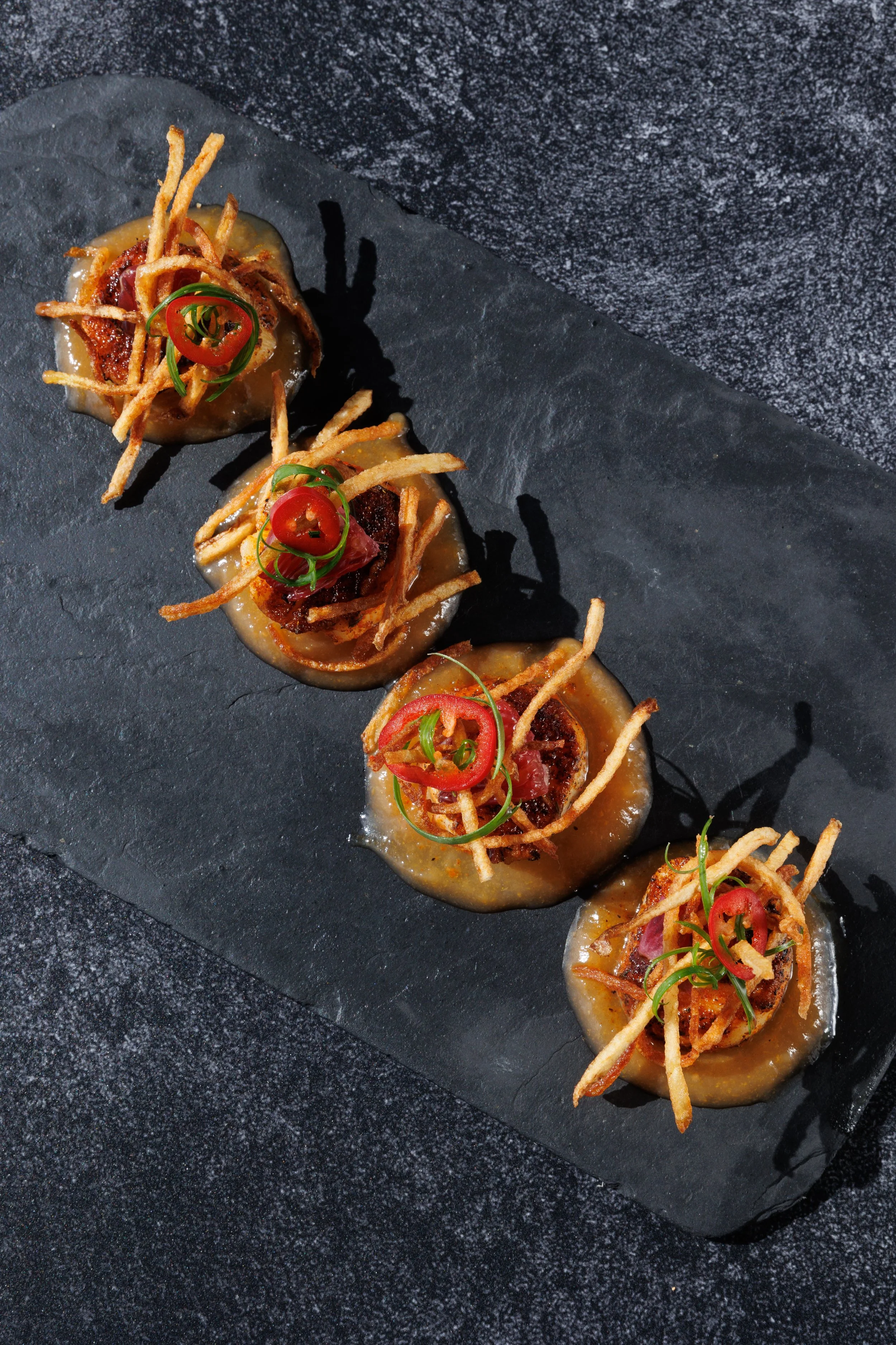 Line of bite-sized appetizers on a black slate serving platter, each topped with crispy fried strips, sliced red chili, and green garnish.