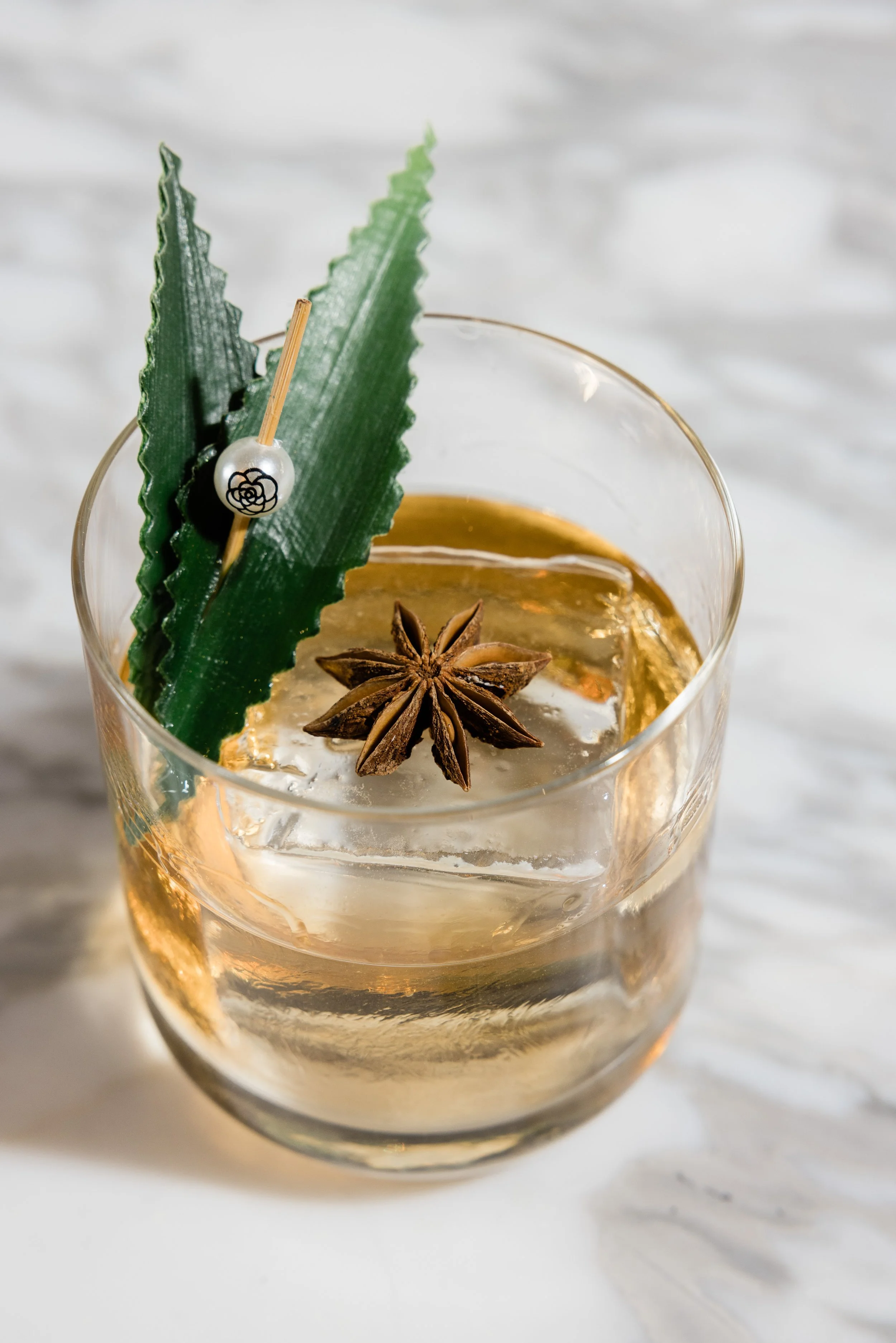 Close-up of a cocktail in a glass with star anise, a green leaf, and a decorative pearl with a flower design.