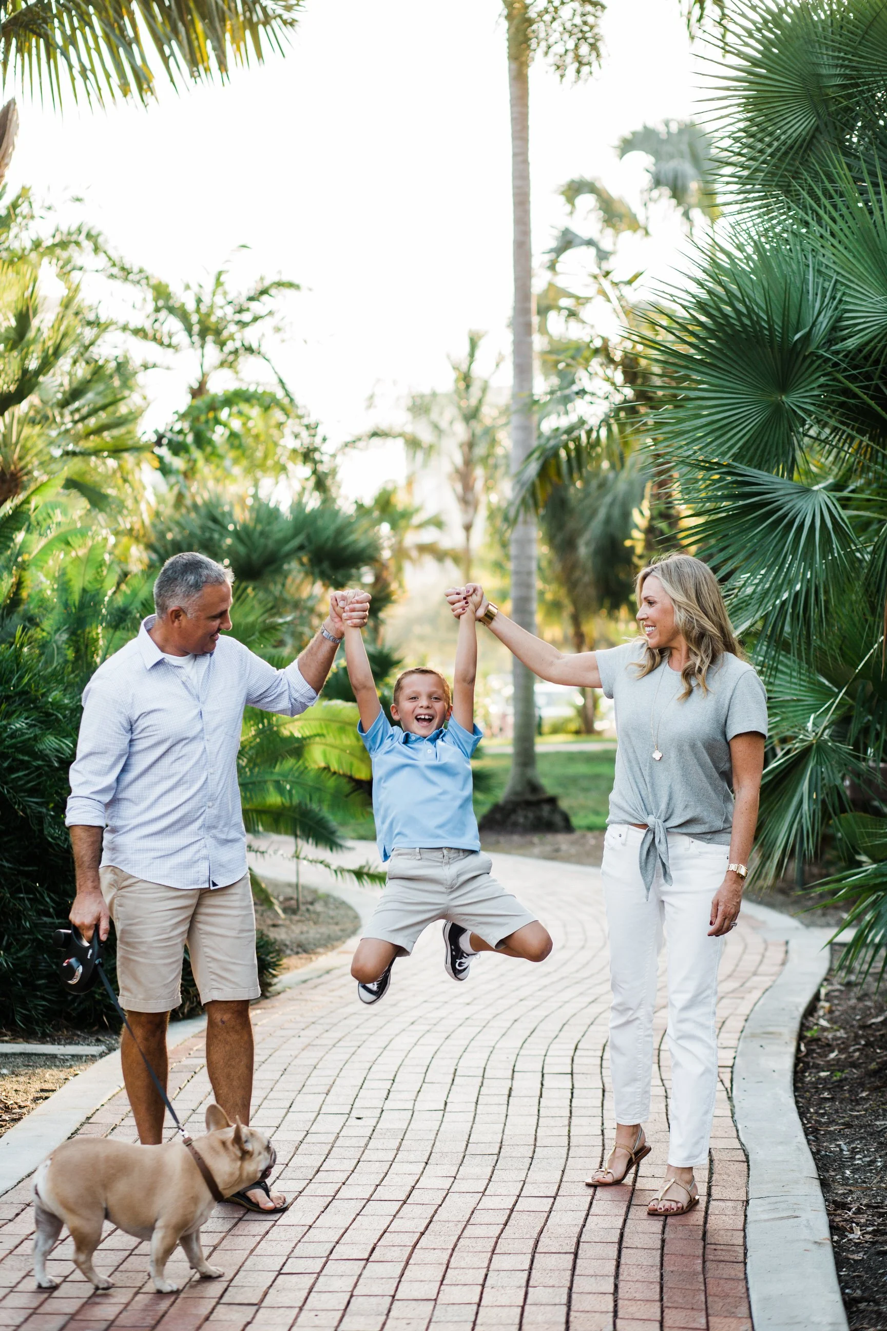 A happy boy is jumping in the air between his grandparents, who are holding his hands. The scene is outdoors on a brick pathway surrounded by lush greenery and palm trees. A woman and man are smiling at the boy, and a dog is on a leash nearby.