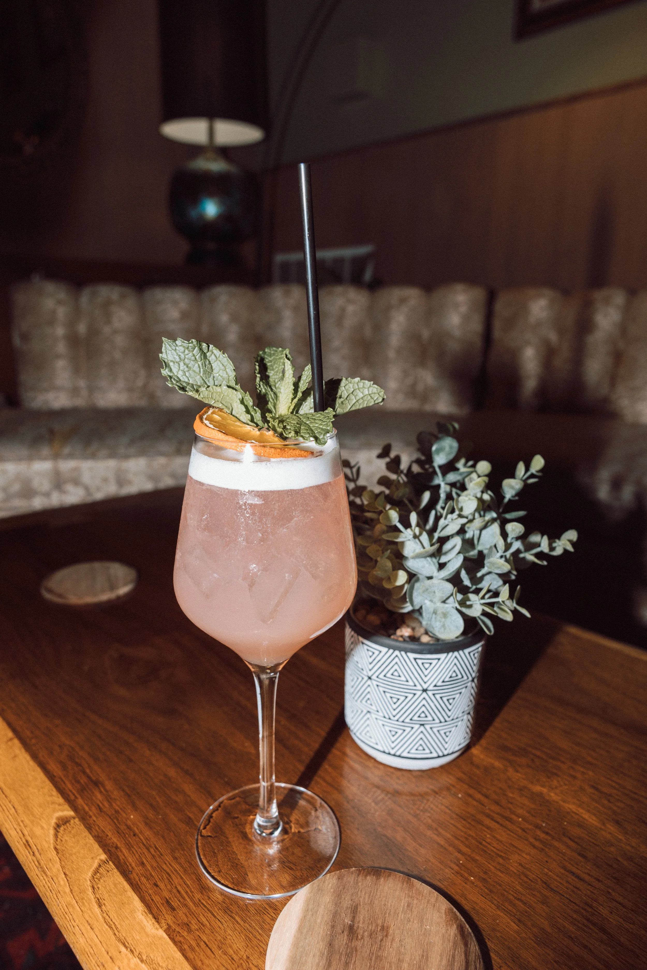 A pink cocktail garnished with mint leaves, an orange slice, and a black straw on a wooden table, with a potted plant nearby and a blurred background featuring a sofa and a lamp.