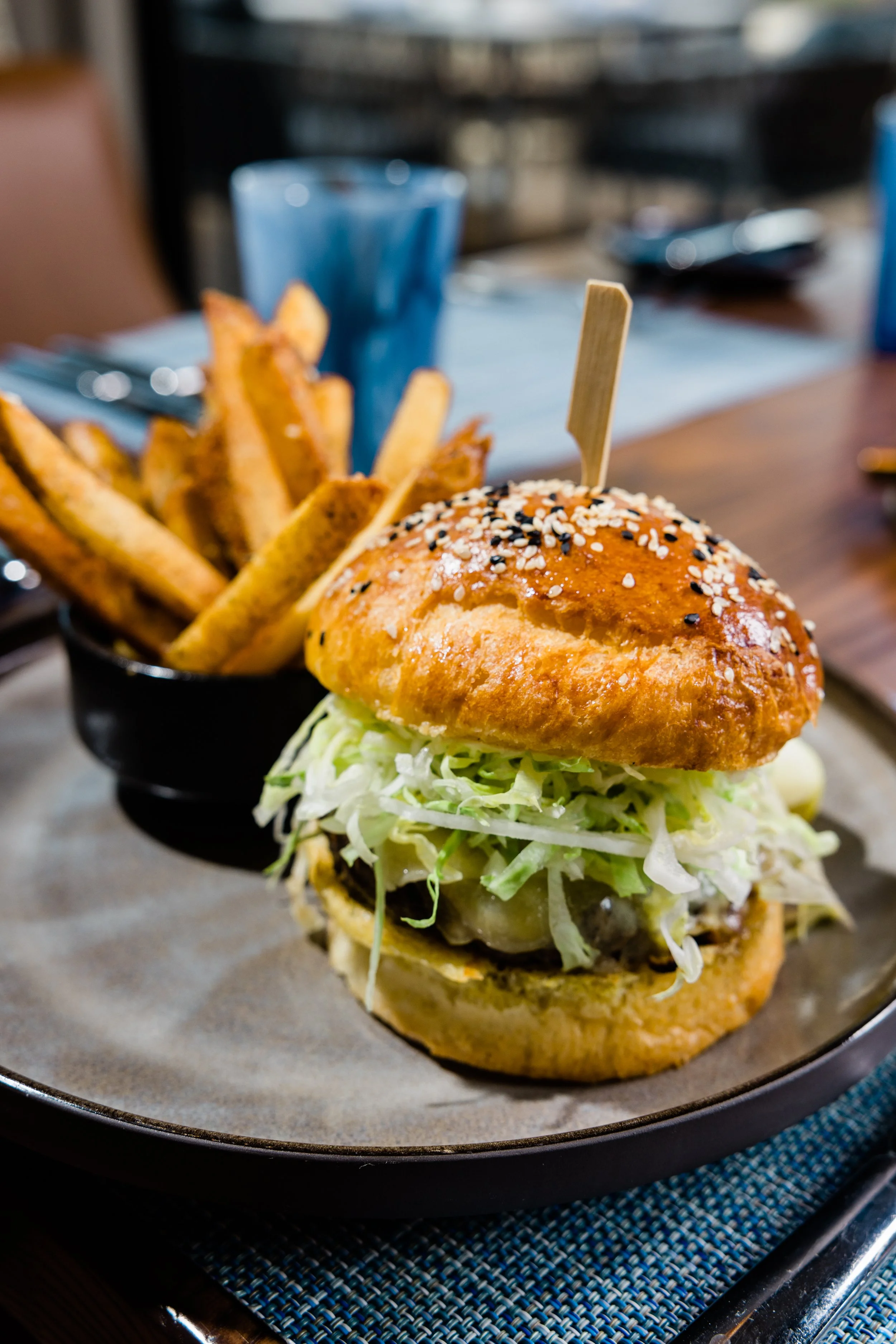 A burger with shredded lettuce and a sesame seed bun on a round metal tray, with a side of French fries in a black cup, on a wooden table.