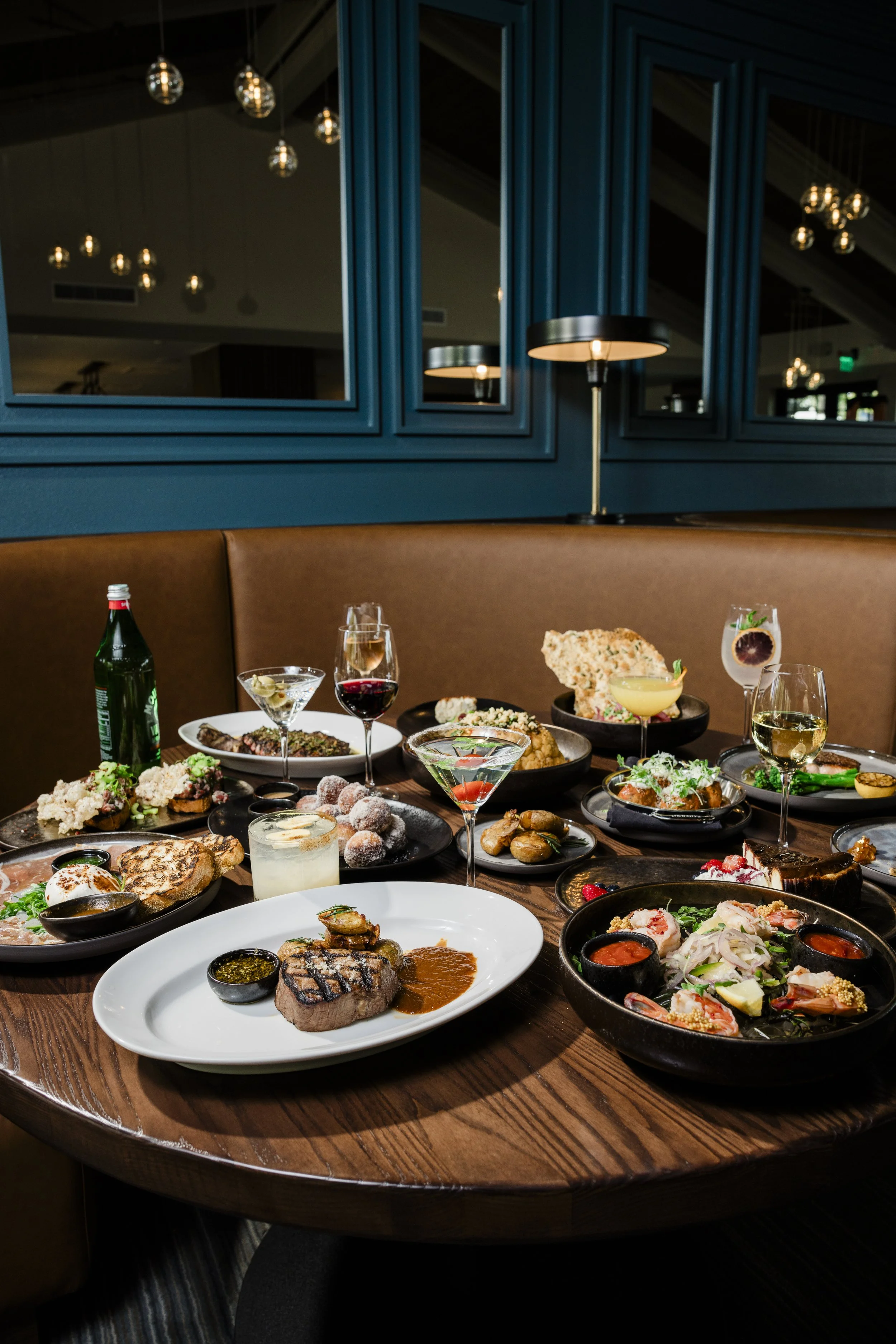 A table filled with various dishes including steaks, salads, appetizers, and drinks in a restaurant with blue-painted walls and modern hanging lights.
