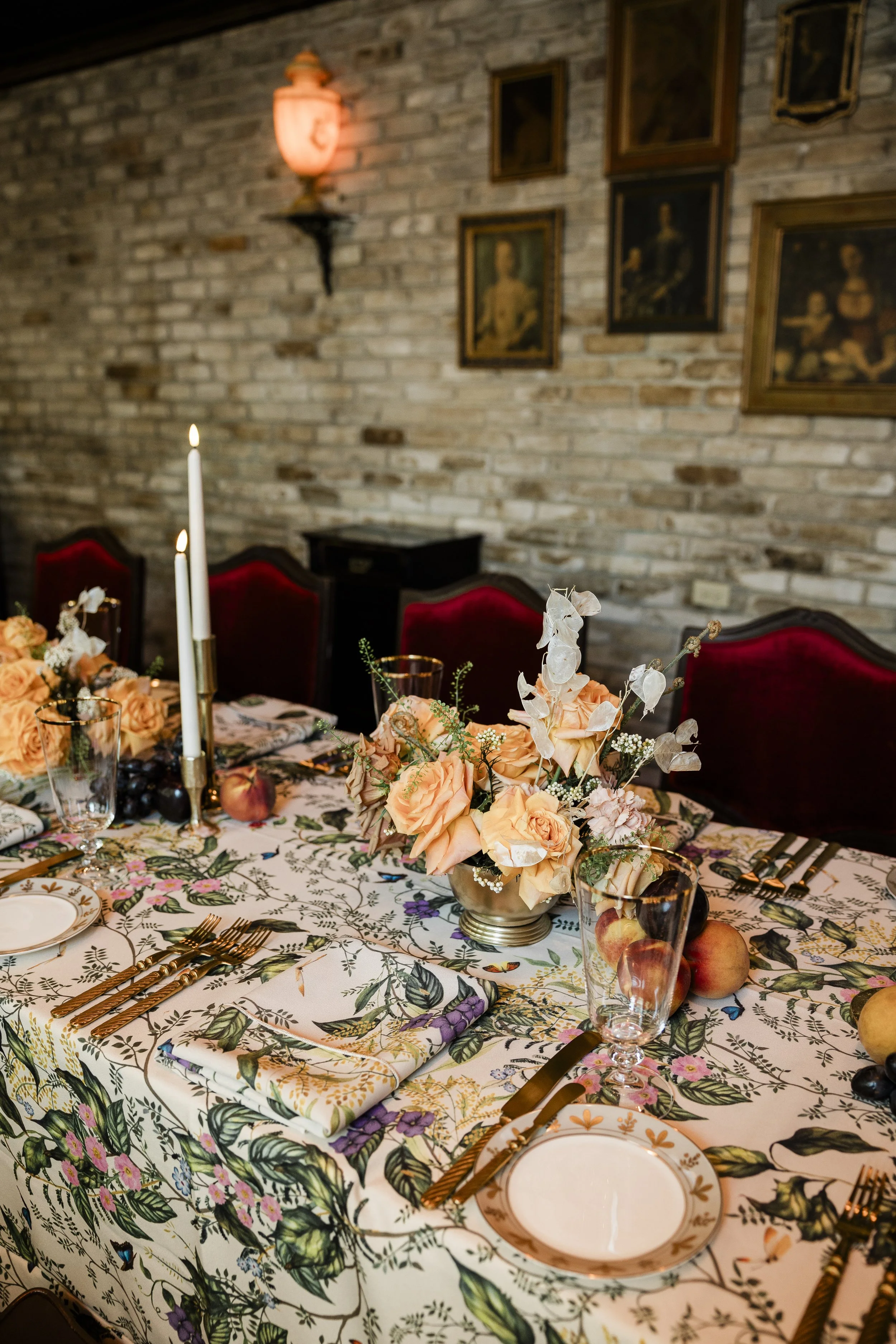 A decorated table with floral-patterned tablecloth, peach roses centerpiece, white candles, gold utensils, and fine plates, set against a brick wall with framed artwork and warm lighting.