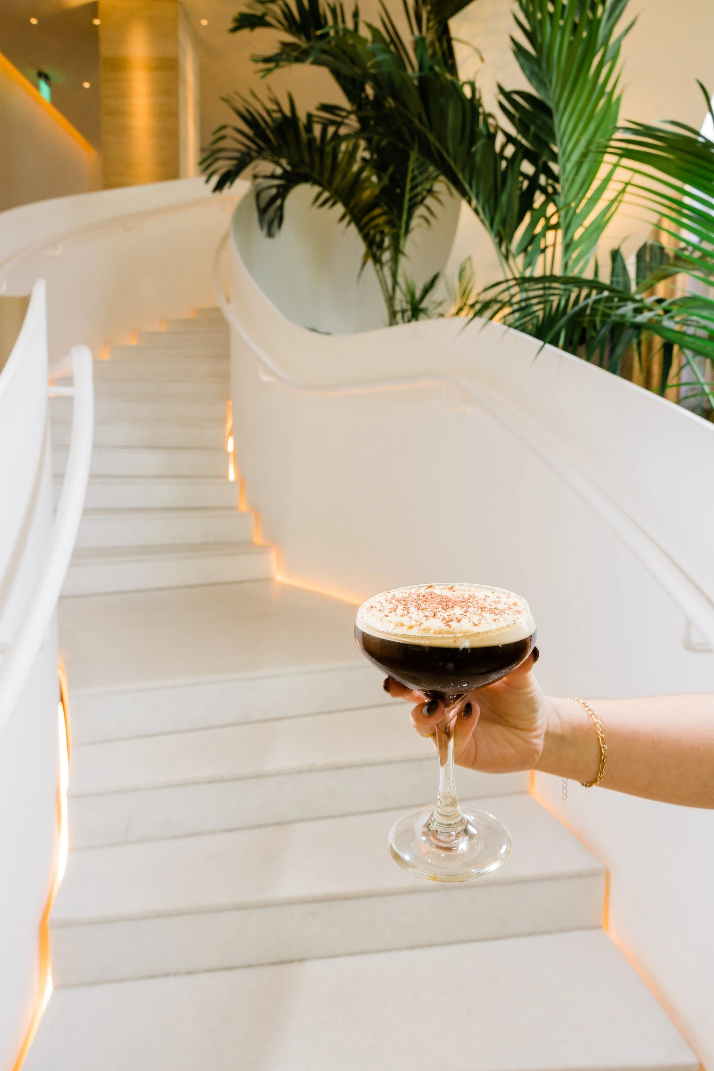 A hand holding a glass of dessert coffee with foam and chocolate sprinkles in front of white staircase with plants in the background.