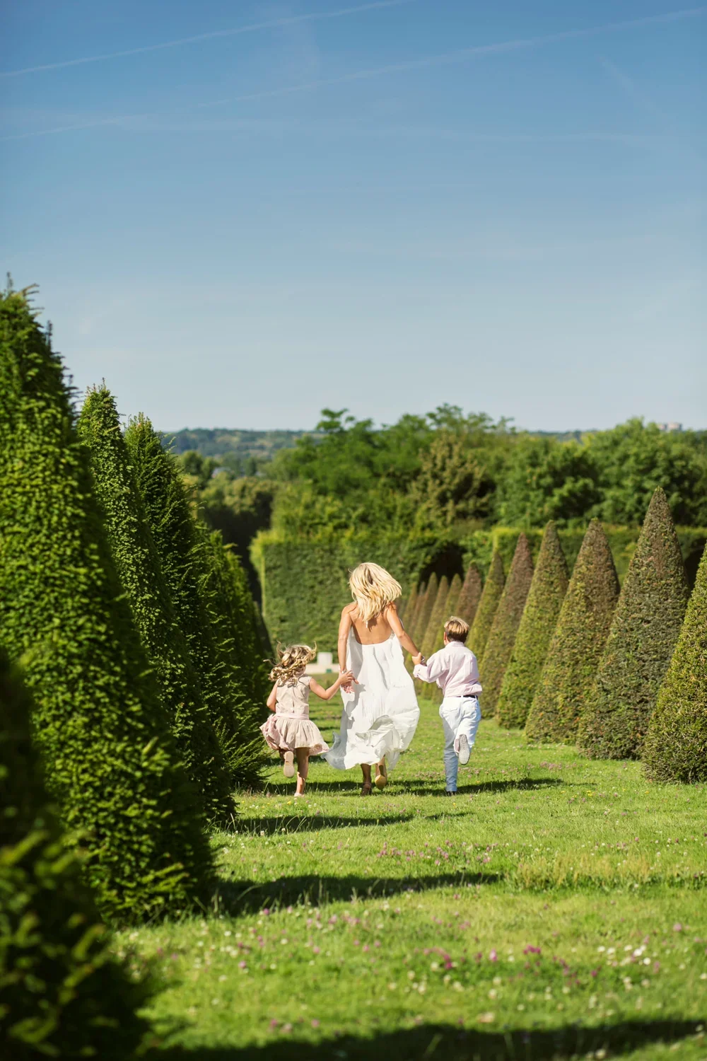 A woman and two children running through a well-manicured garden of Versailles with tall, cone-shaped shrubs on either side, under a clear blue sky in France.