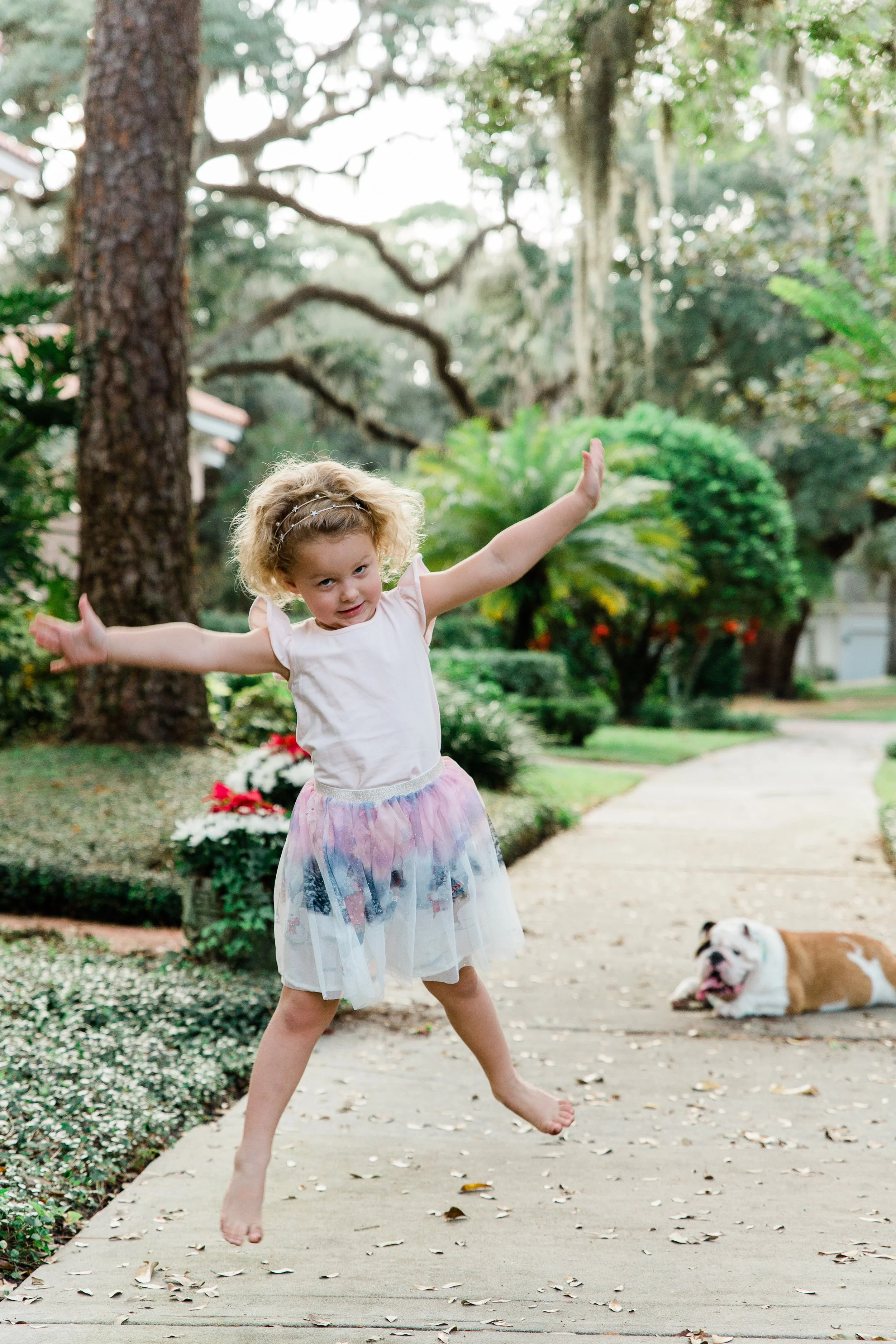 little girl in white shirt and ombre skirt jumping in the air with arms outstretched. Bulldog watches in the background.