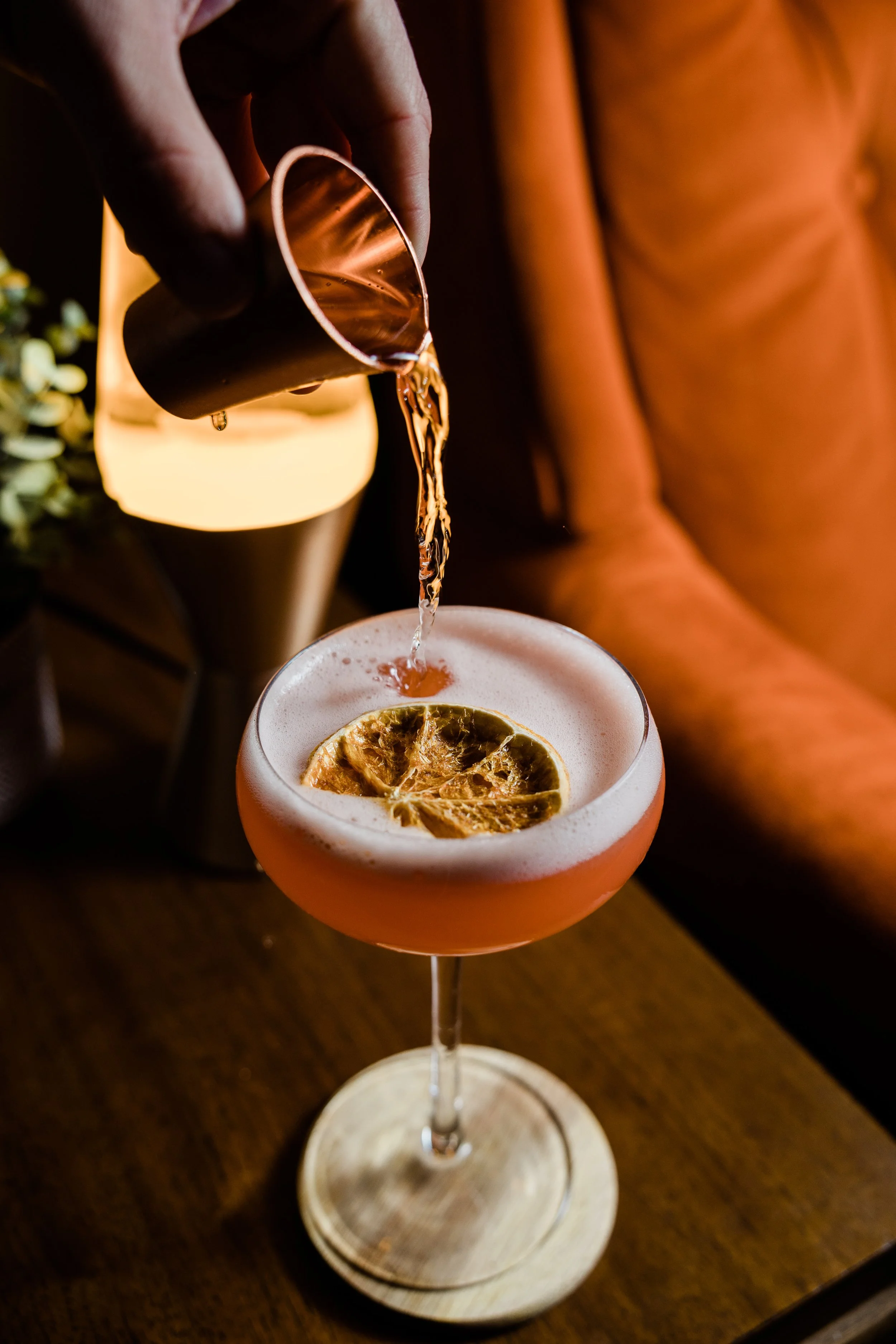 A person pouring a drink into a cocktail glass with a dried lemon slice garnish on top, sitting on a wooden table.