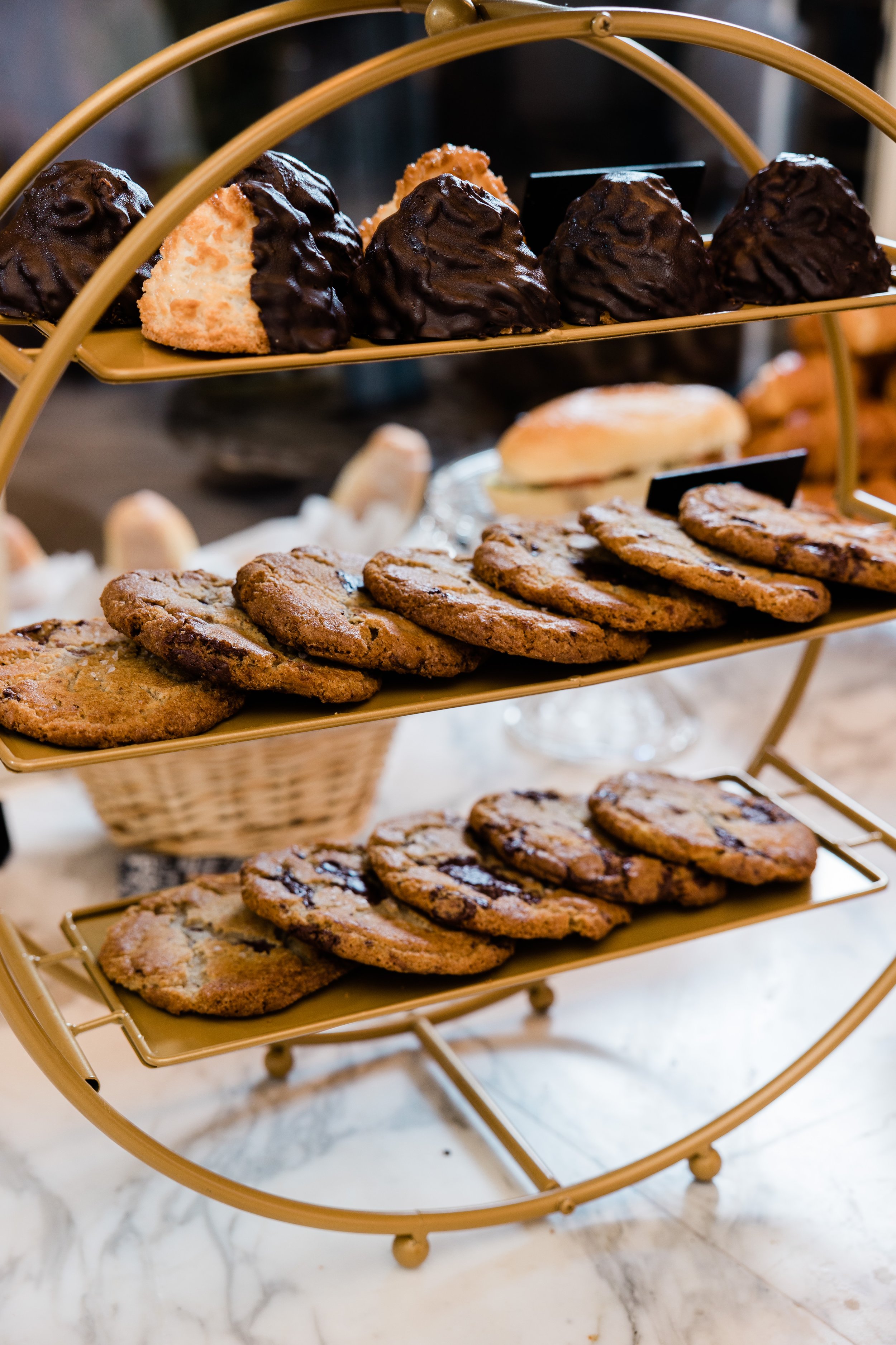 A three-tiered golden serving stand with chocolate-covered cookies on the top tier and chocolate chip cookies on the middle and bottom tiers, placed on a marble surface.